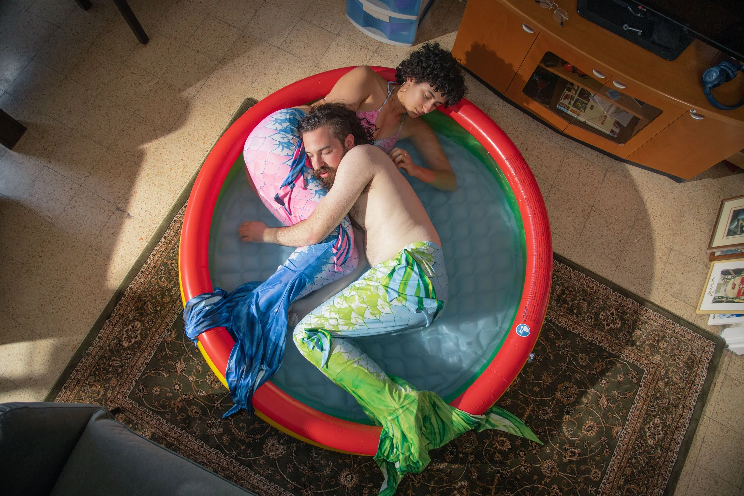 Two people laying in an inflatable pool, one with curly hair and the other with short gray hair, both appears to be sleeping inside a room with TV and furniture.