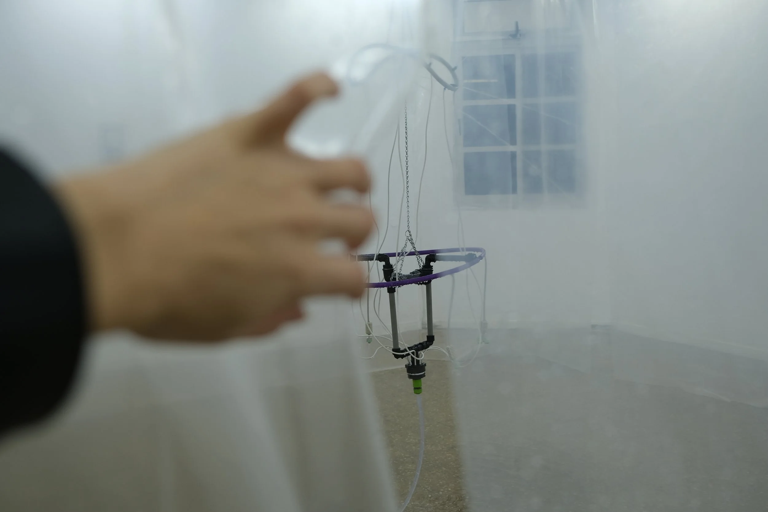 A blurred hand reaching towards a small robotic or mechanical device on a stand with various wires and components, seen through a semi-transparent surface.