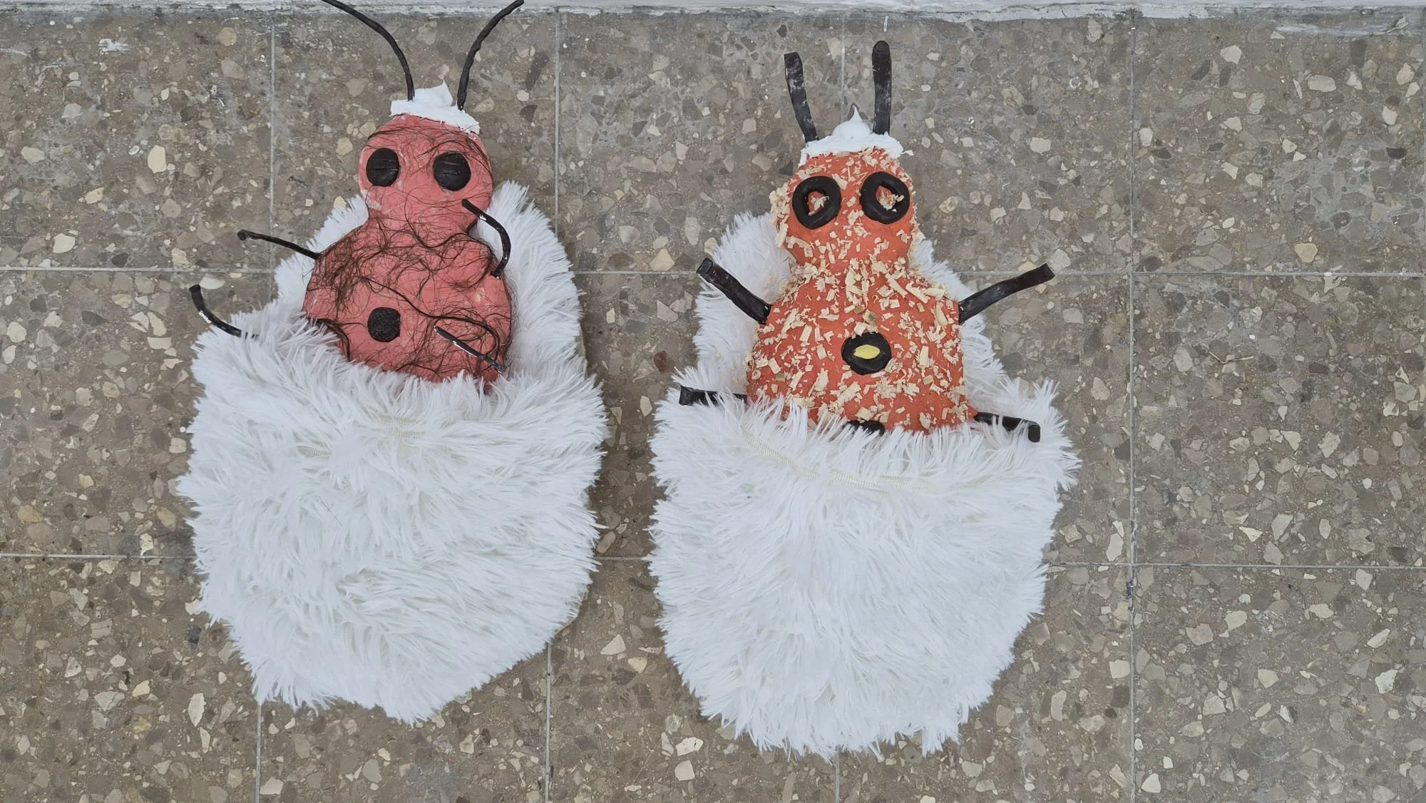 Two handmade paper mache ladybug and firefly crafts with white feathery wings on a tiled floor.