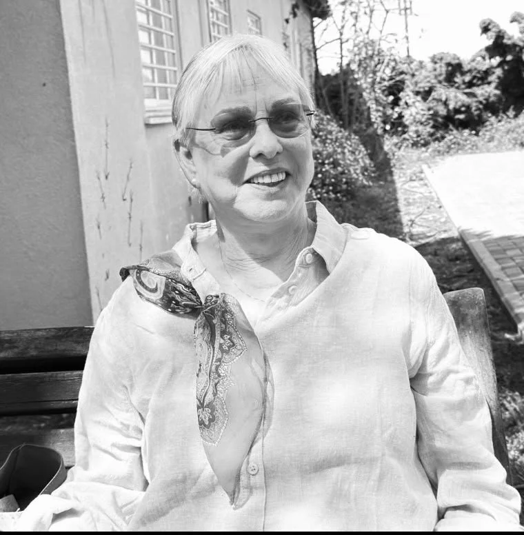 A smiling elderly woman with short white hair, wearing glasses, a light-colored button-up shirt, and a patterned scarf tied around her neck, sitting outdoors on a wooden bench.