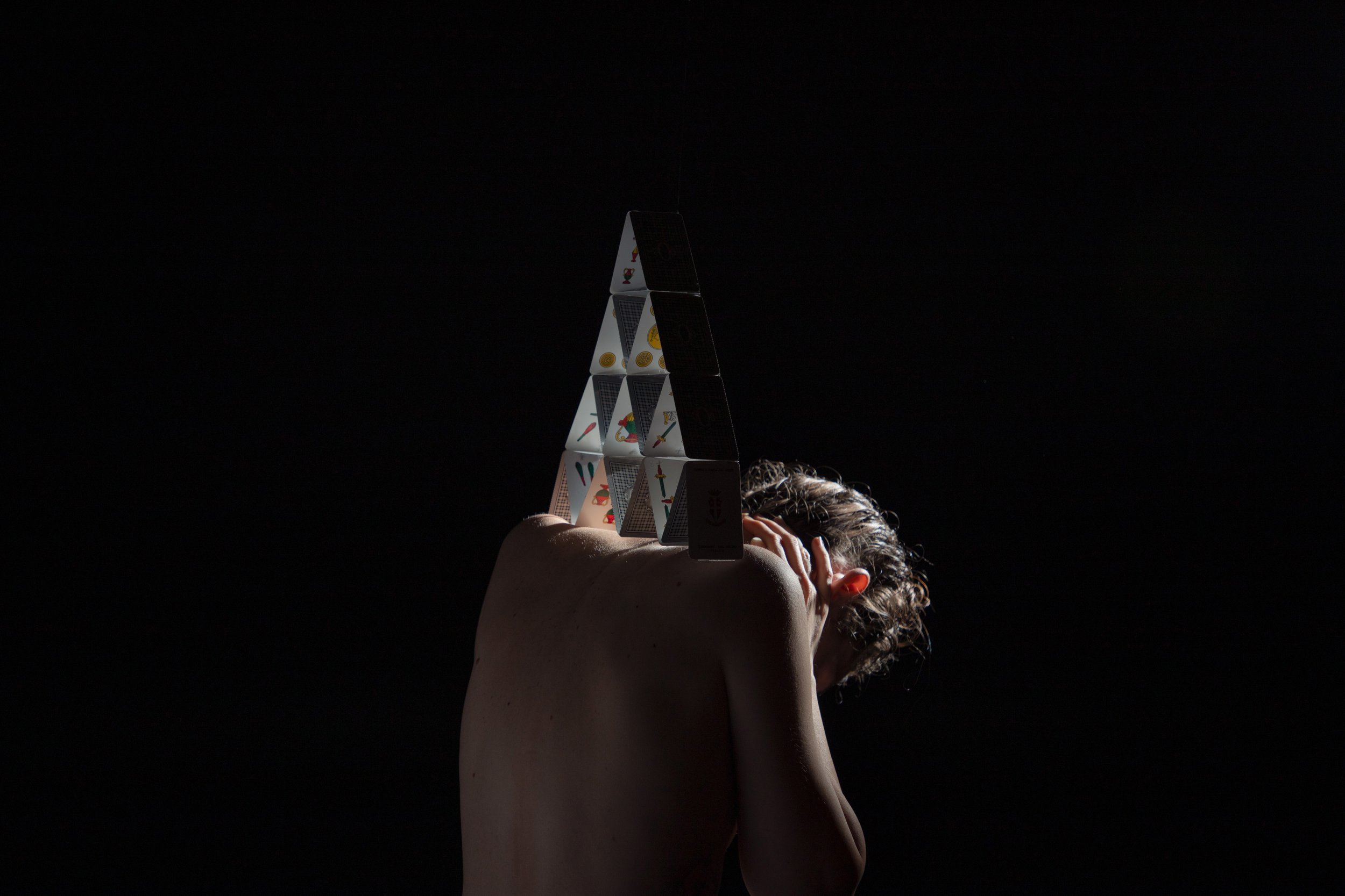 A person with light skin and curly hair is lying down with their head tilted back, and their arm is raised with a hand on their forehead. Their face and body are mostly in shadow, with a dark background. A tower of standard playing cards is balanced on their arm, with some cards showing symbols and patterns.