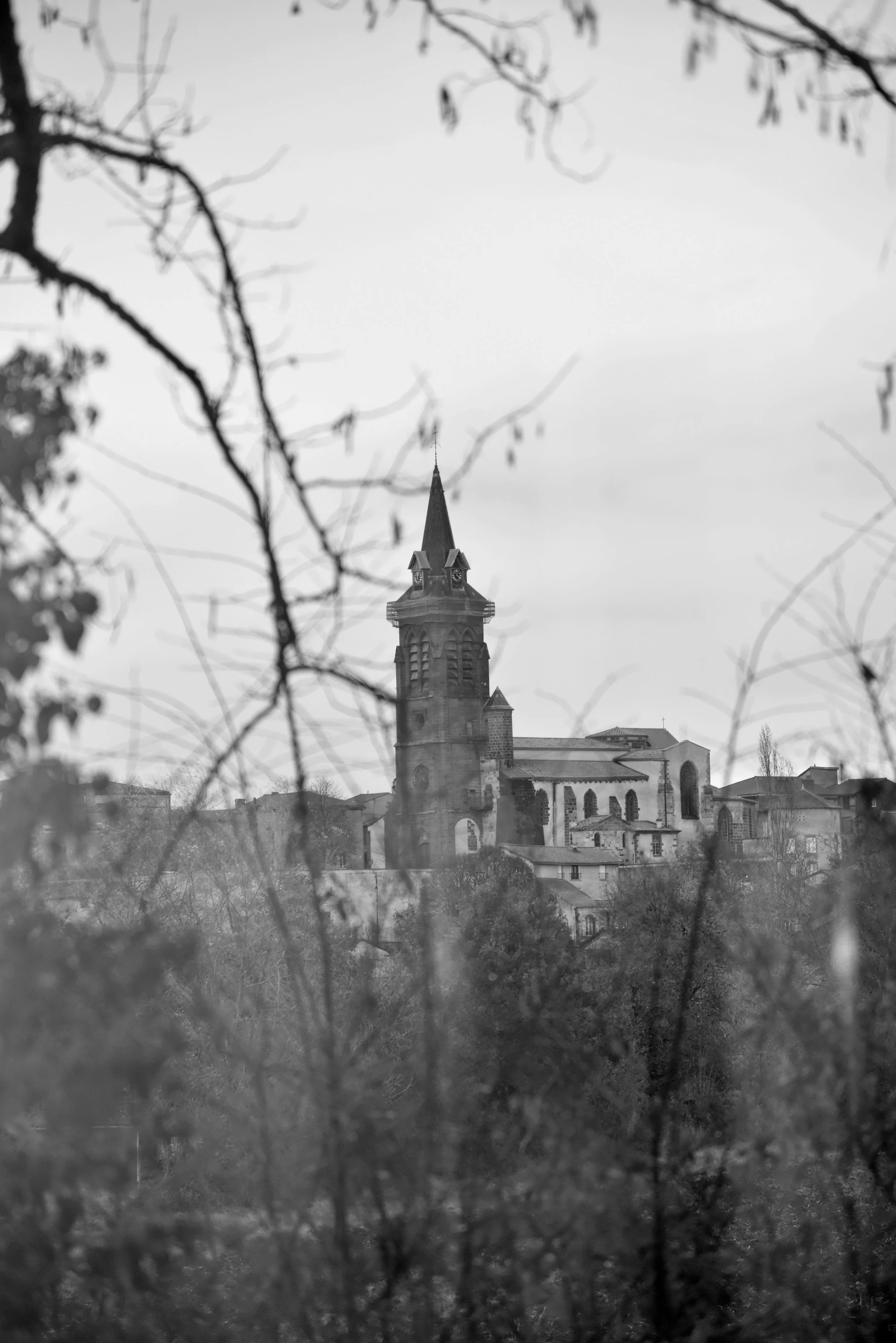A black-and-white photograph of a church with a tall steeple, seen through tree branches and foliage.