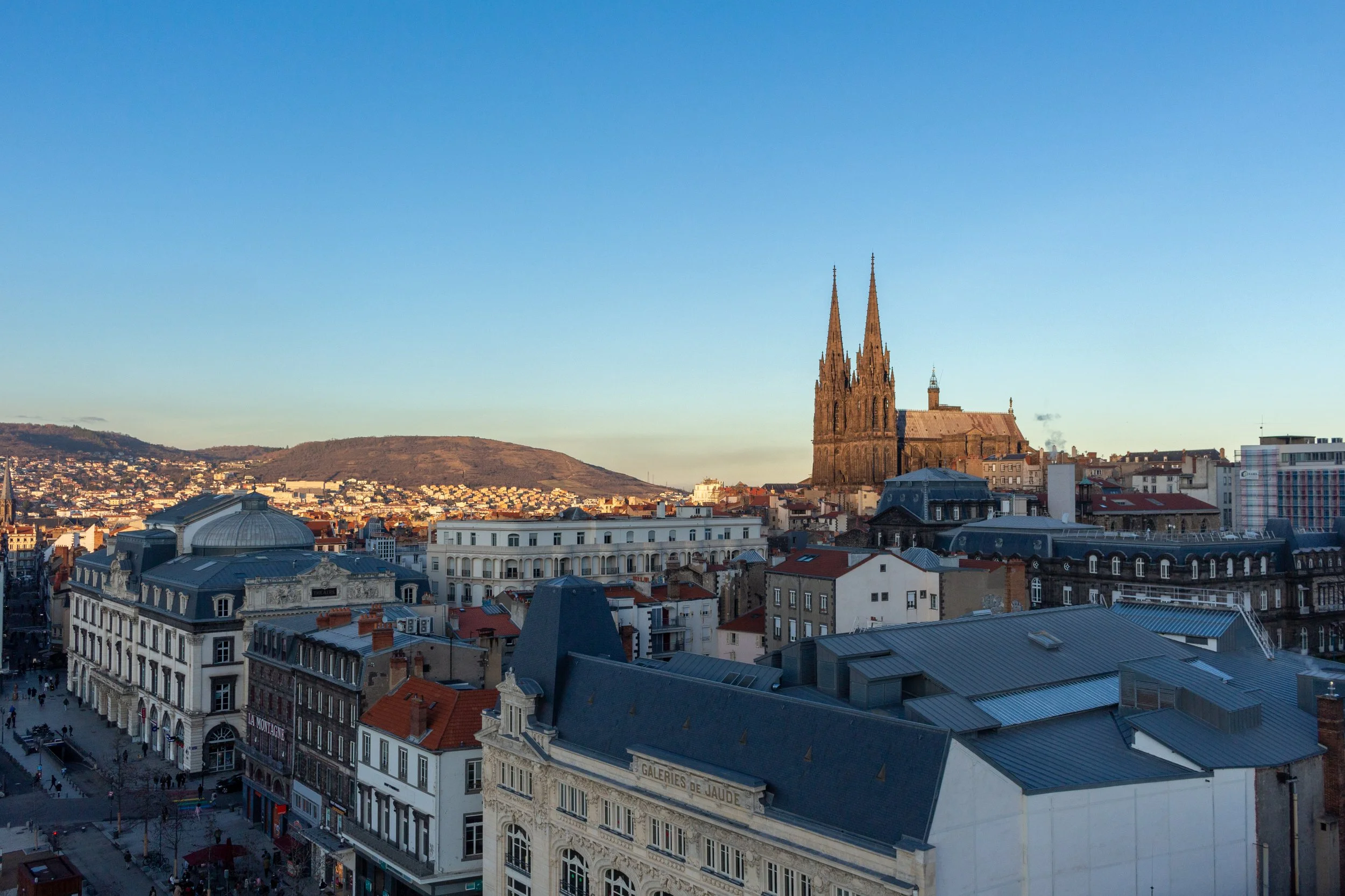 Cityscape of a European city with a large Gothic cathedral, surrounding buildings, and a hill in the background under a clear blue sky.