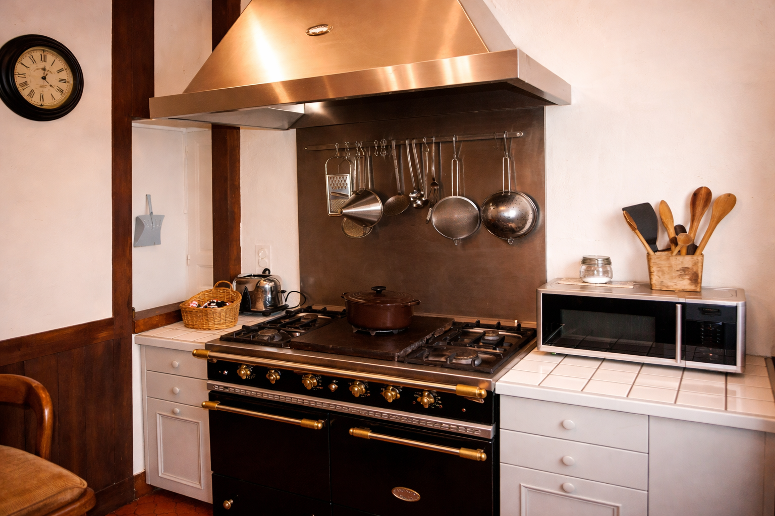 Une cuisine avec une cuisinière noire, un micro-ondes posé sur un plan de travail carrelé de blanc, et des ustensiles de cuisine suspendus au-dessus de la cuisinière, dont une râpe, des louches et une passoire. Une horloge est fixée au mur, et un pan