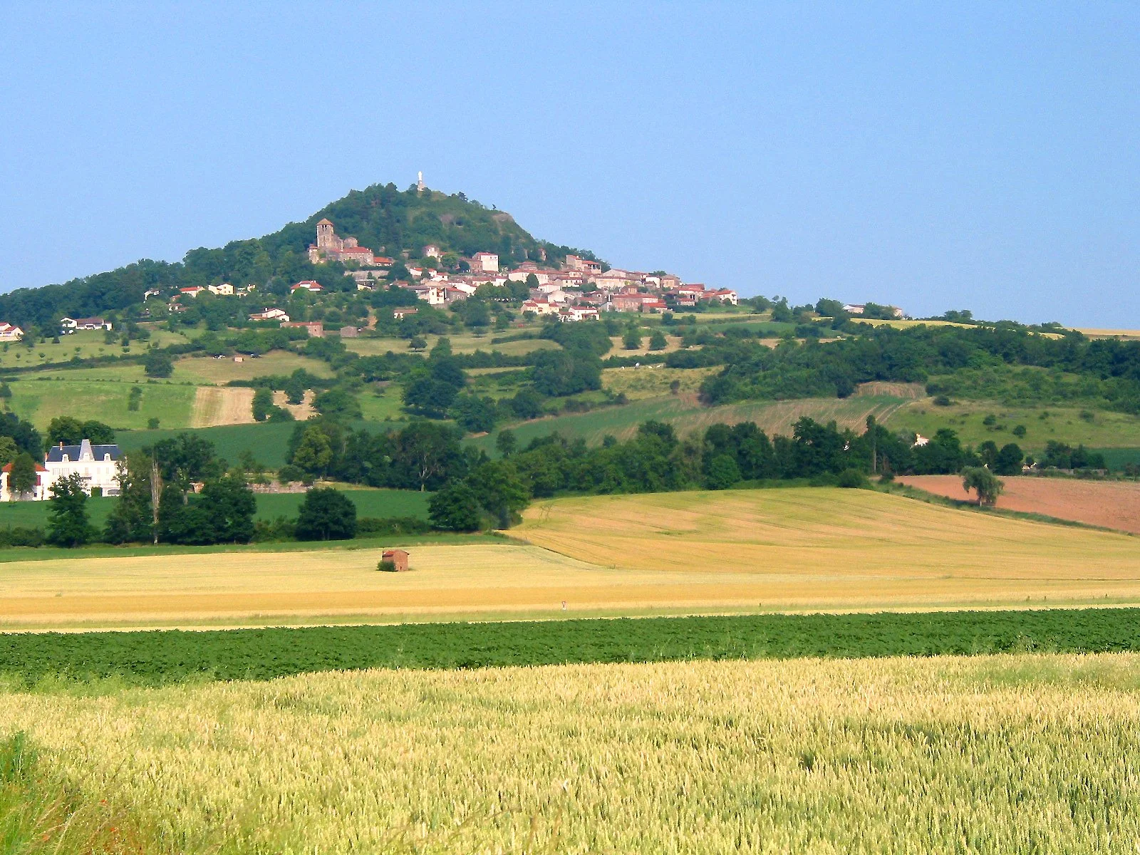 Scenic view of a hillside village with houses and a church, overlooking cultivated fields and green landscape under a blue sky.