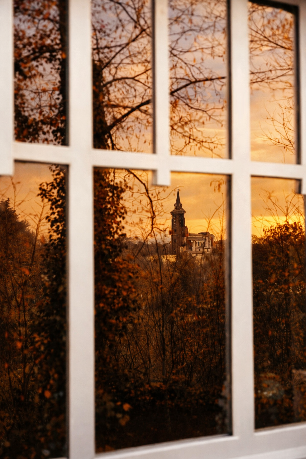 Sunset view through a window with white grid frames, showing a church steeple surrounded by trees with autumn leaves.
