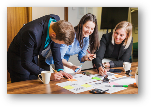 Tres personas trabajando juntas en una mesa con documentos, marcadores, una calculadora y tazas, sonriendo y colaborando.
