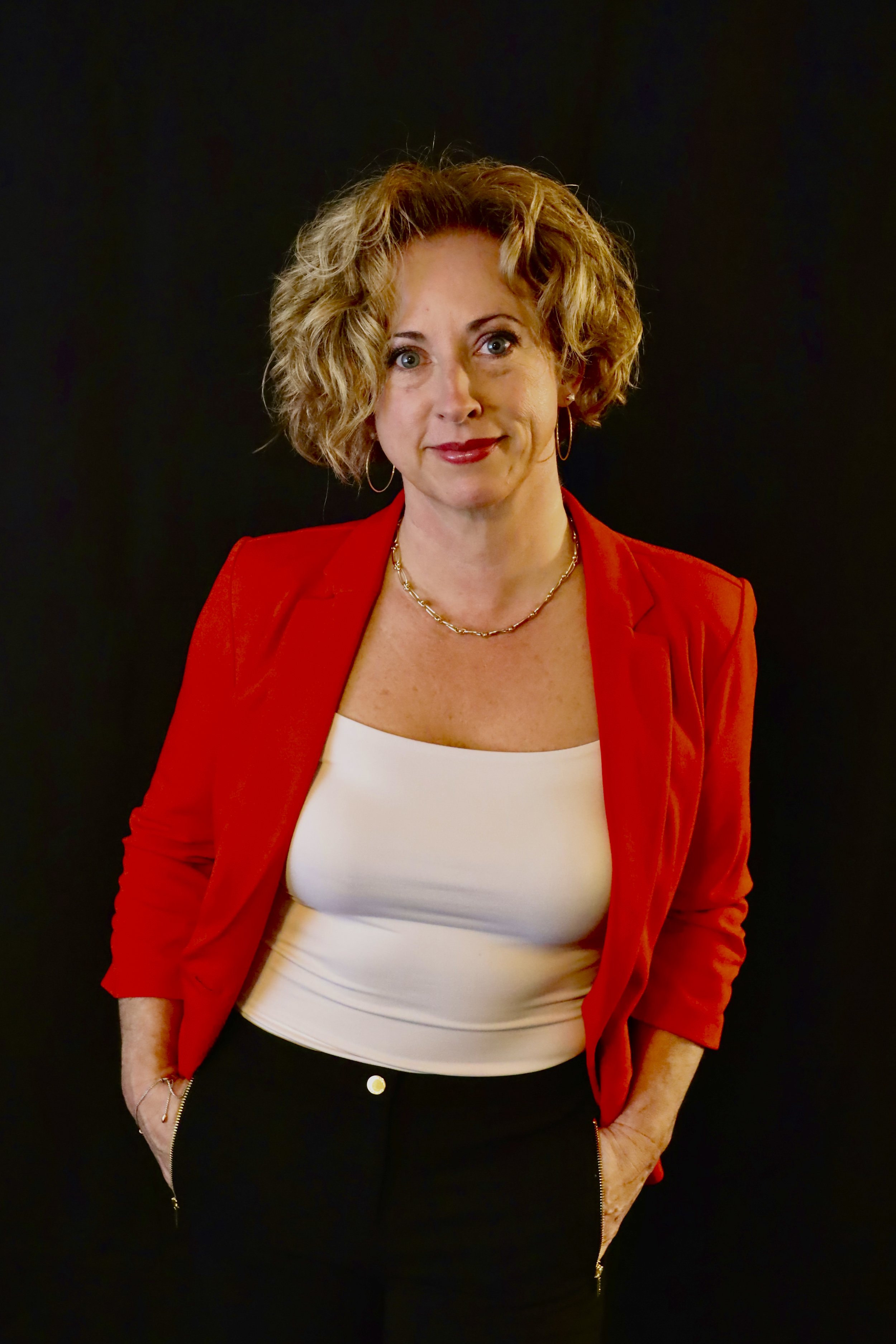 A woman with curly blonde hair wearing a red blazer, white top, and black pants, standing against a black background.