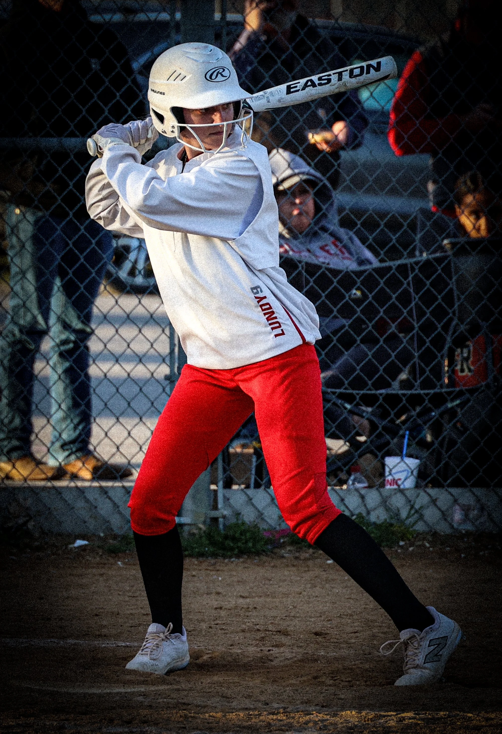 A young baseball player wearing a white helmet, white jersey, red pants, and black socks stands in a batting stance with a baseball bat at a baseball field.