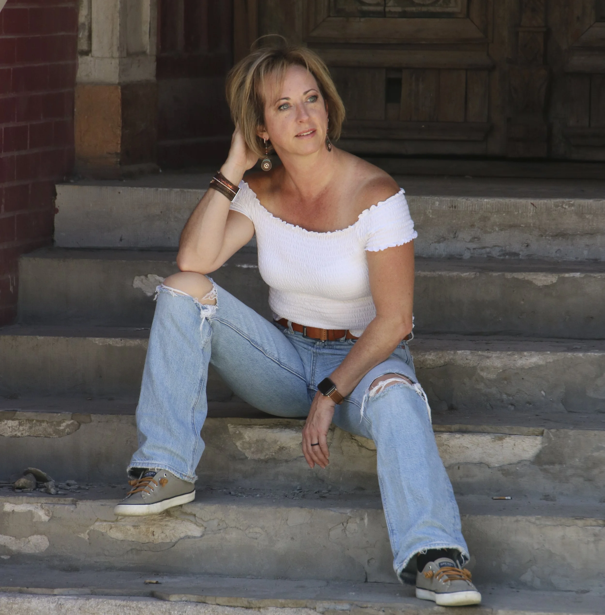 A woman with short light brown hair sitting on concrete steps outside a building, wearing a white off-the-shoulder top, ripped jeans, and sneakers. She is resting her left arm on her knee and touching her hair with her right hand.