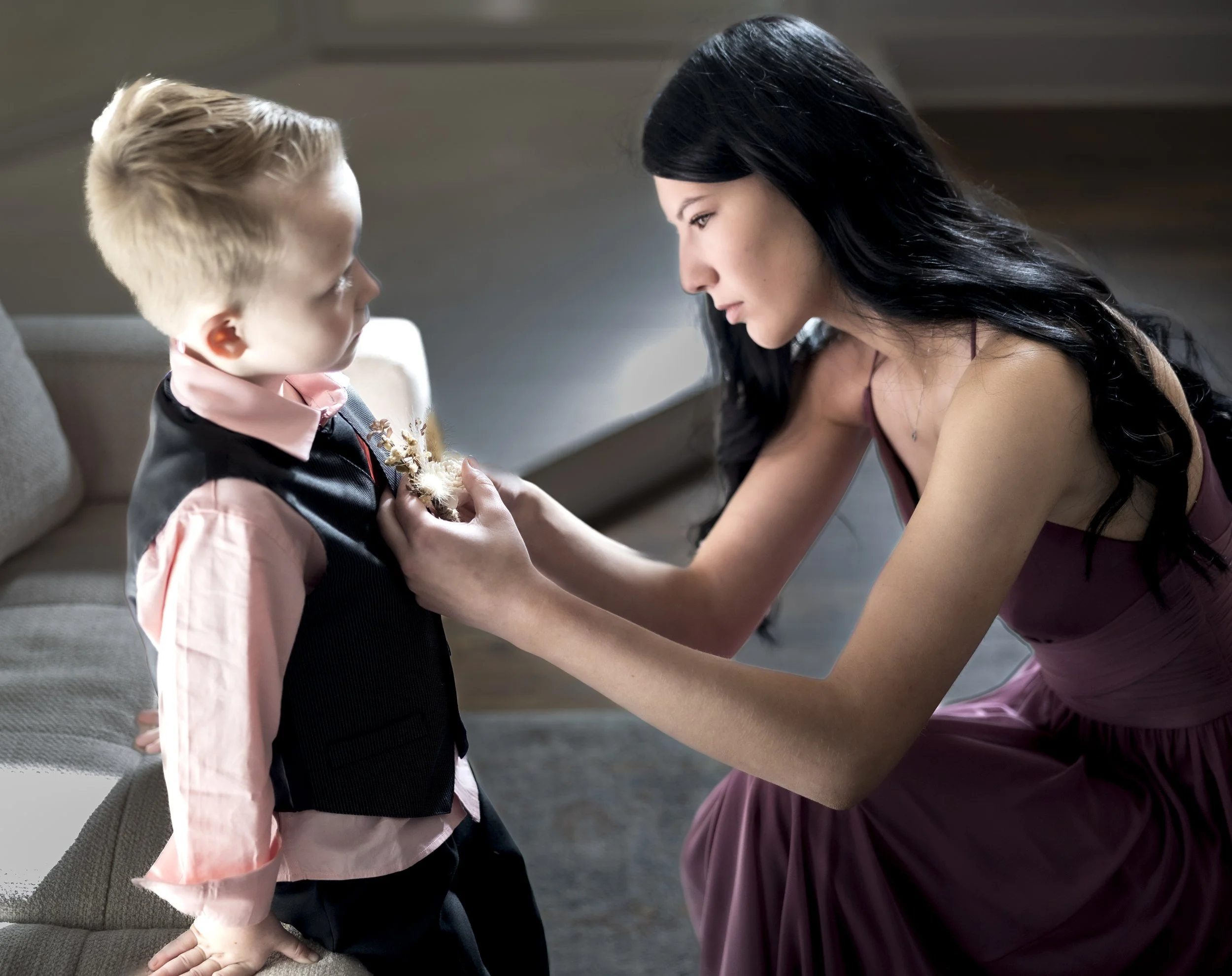 A woman in a mauve dress helps a young boy in a suit adjust a boutonniere on his vest.