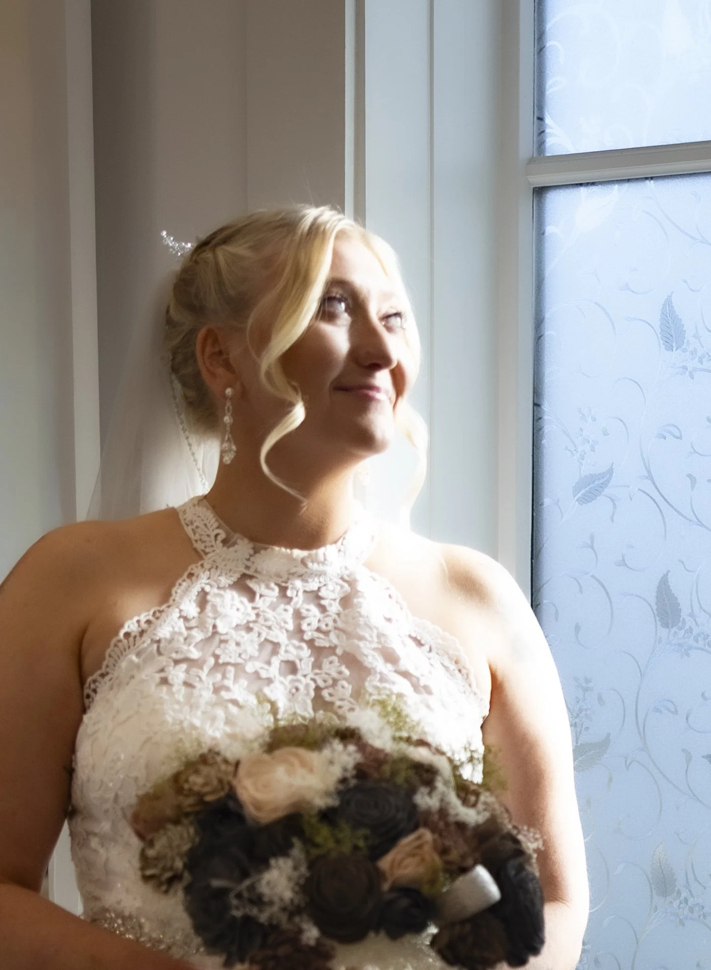 A bride with blonde hair looking up and smiling, holding a bouquet of dark and light flowers, standing near a frosted window with floral patterns, wearing a lace wedding dress and dangling earrings.