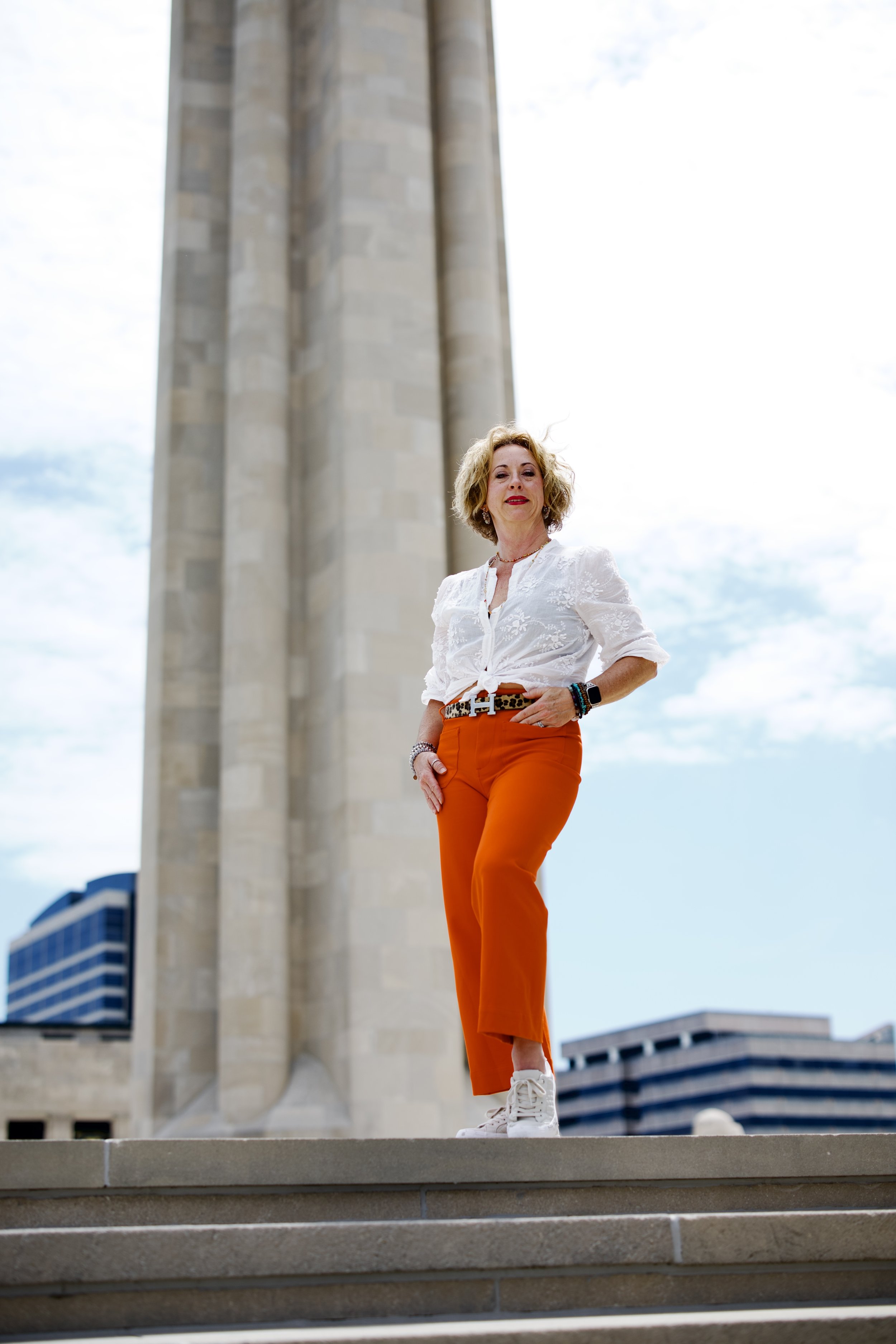 A woman with blonde curly hair standing on outdoor steps in front of a large column with buildings in the background.