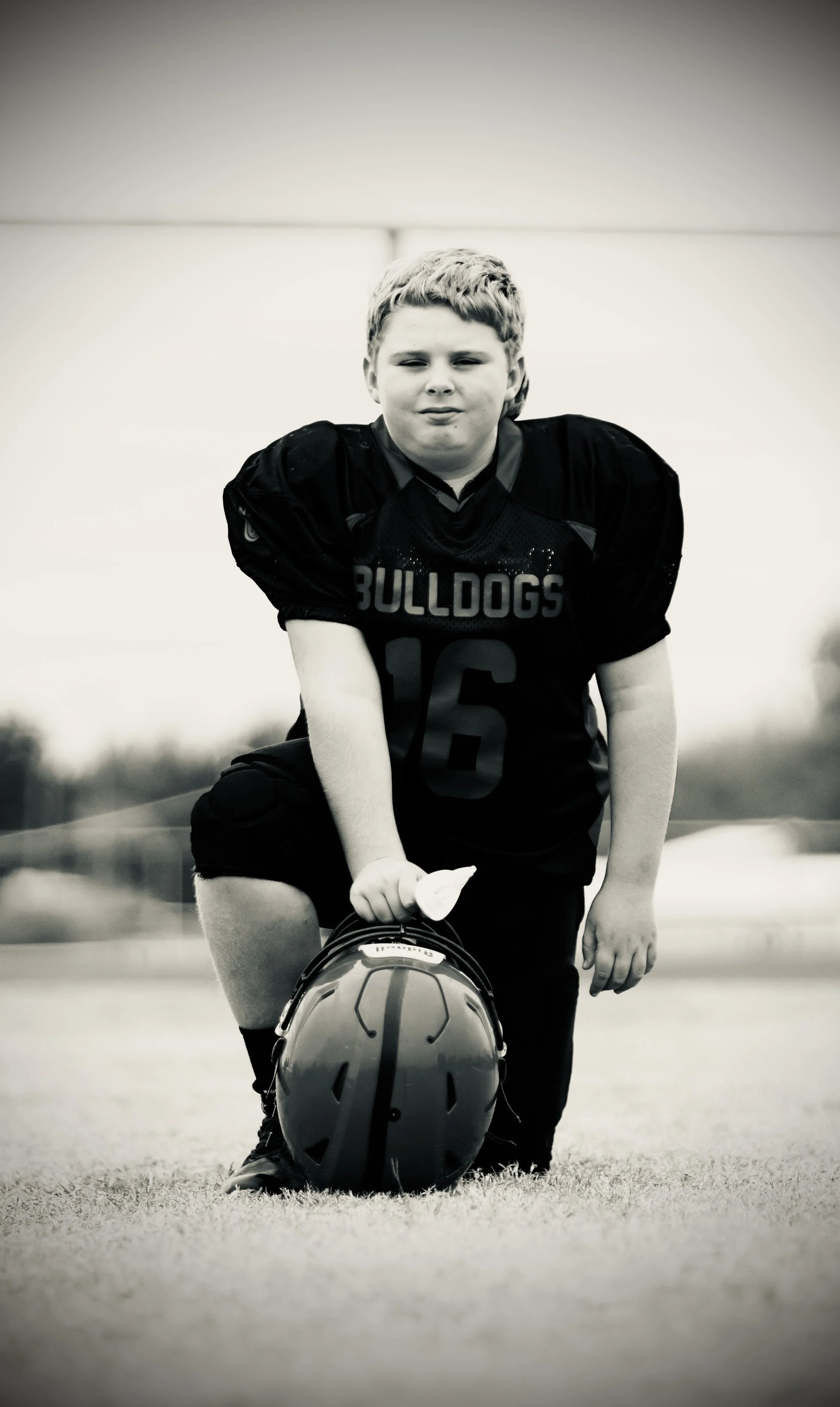 A young boy in a football uniform kneeling on the field, holding a football helmet in front of him.