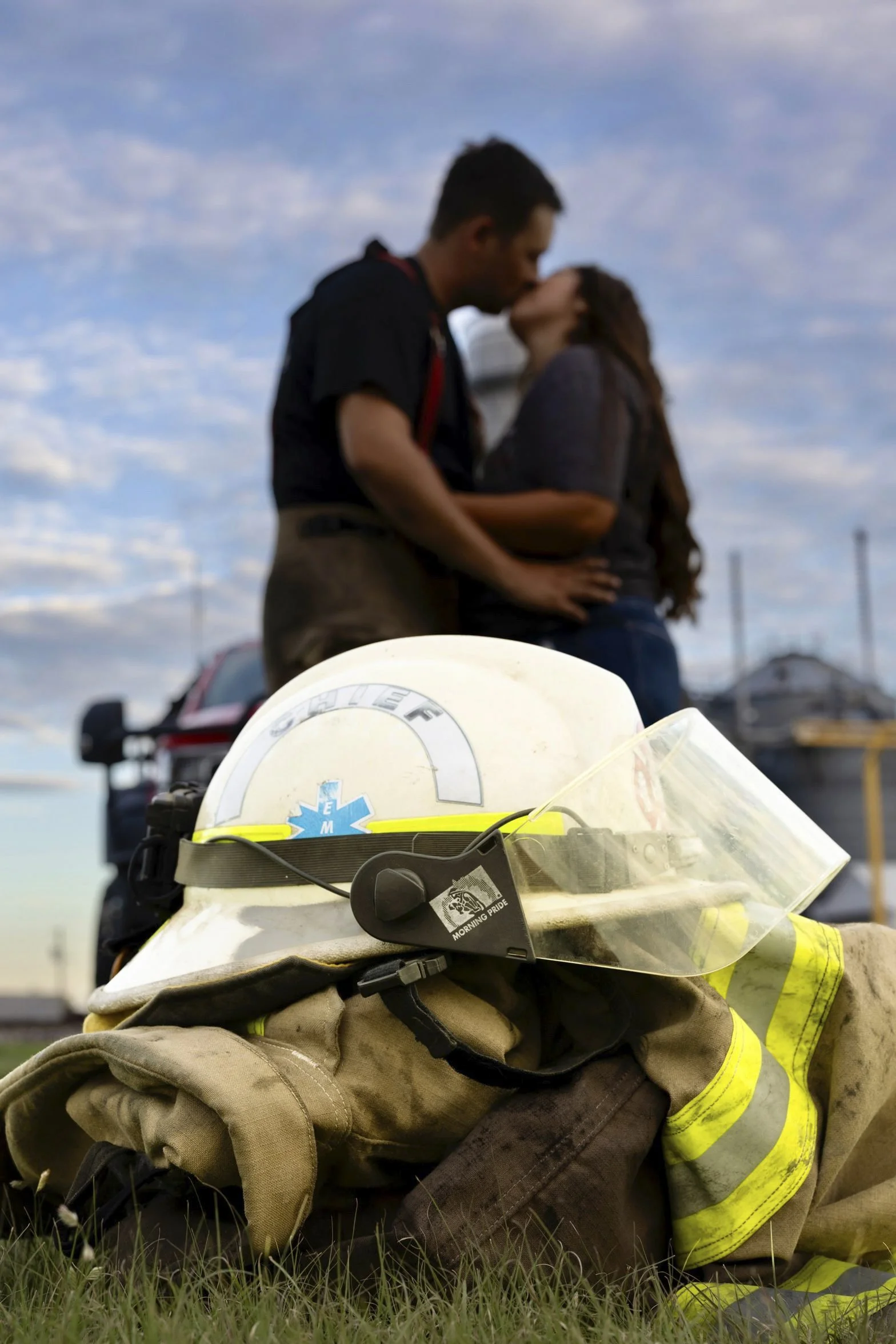 Firefighter helmet and gear in the foreground with an emergency vehicle behind it, and a couple kissing in the background under a partly cloudy sky.