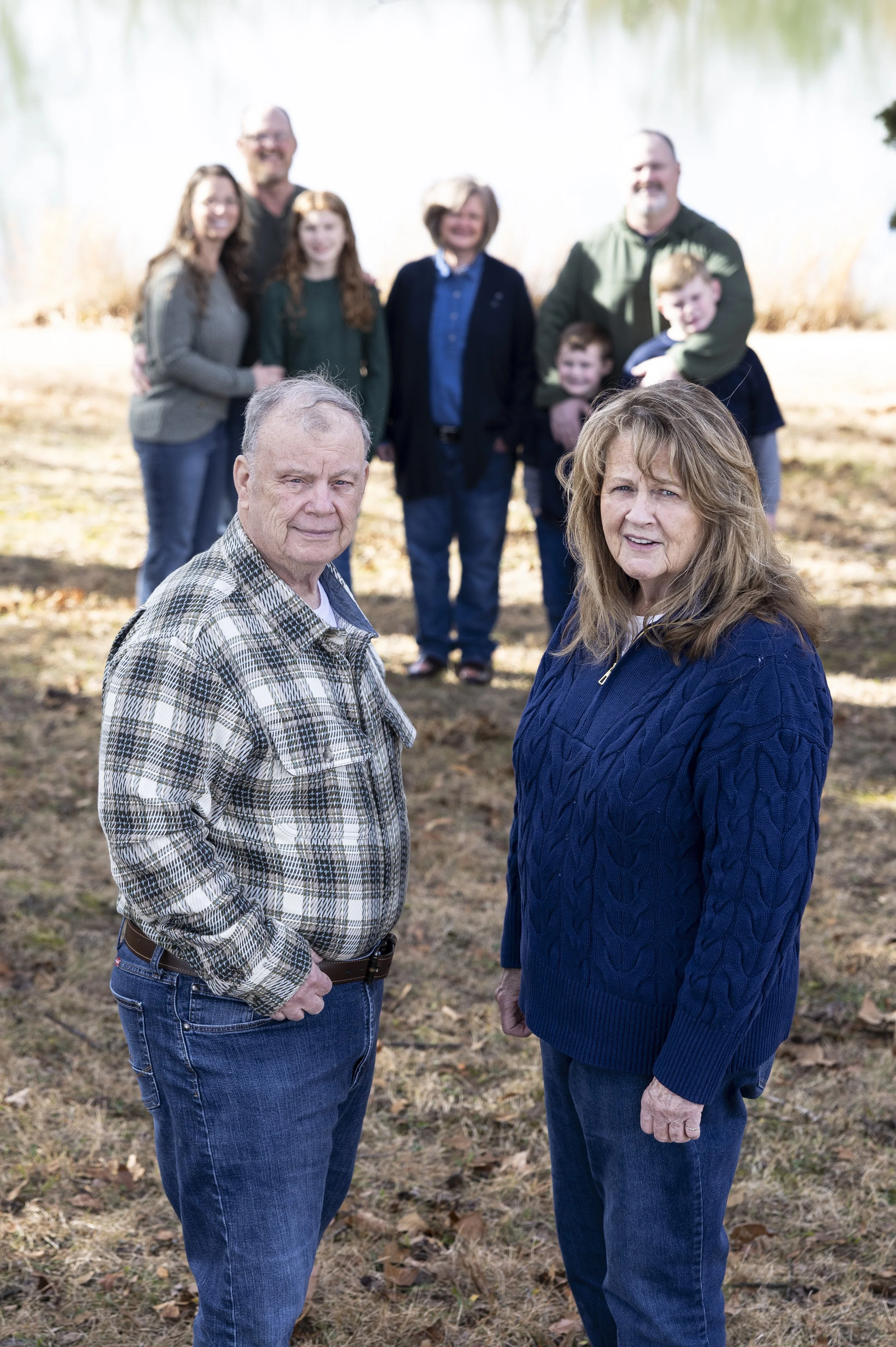 A multi-generational family photo outdoors showing two older adults in the foreground, a man in a plaid shirt and a woman in a navy blue sweater, with a middle-aged woman and man, a girl, and two boys in the background, standing on a grassy area near a lake during daytime.