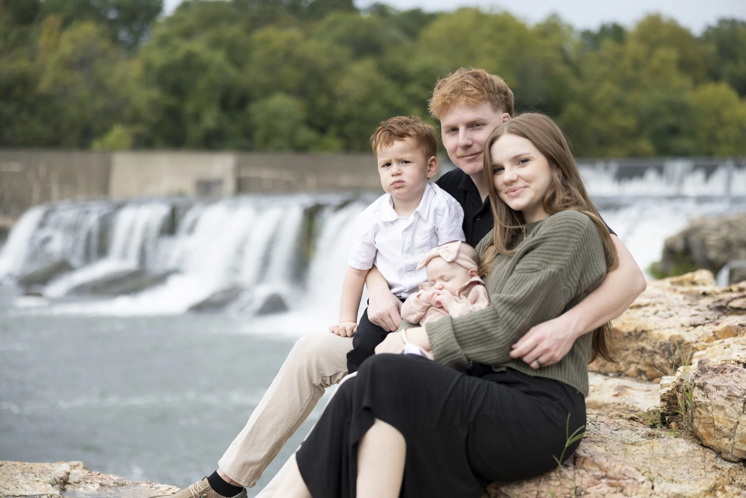 A family of four sitting on rocks near a waterfall, with trees in the background. The father has red hair and is holding a young boy with red hair. The mother has long brown hair and is holding a baby girl in a pink outfit and bow.