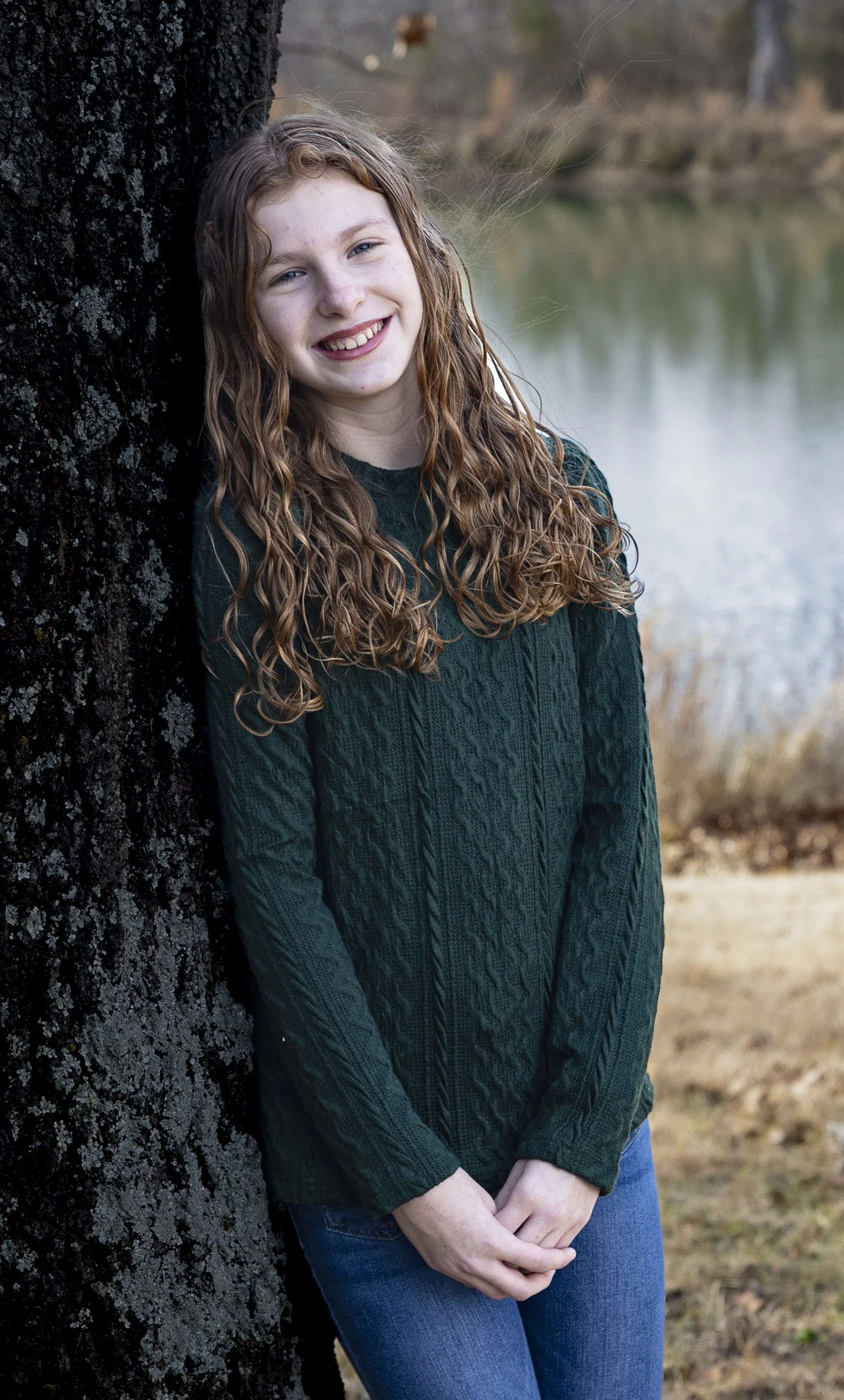 A young woman with curly red hair, smiling, leaning against a tree near a lake in an outdoor setting.