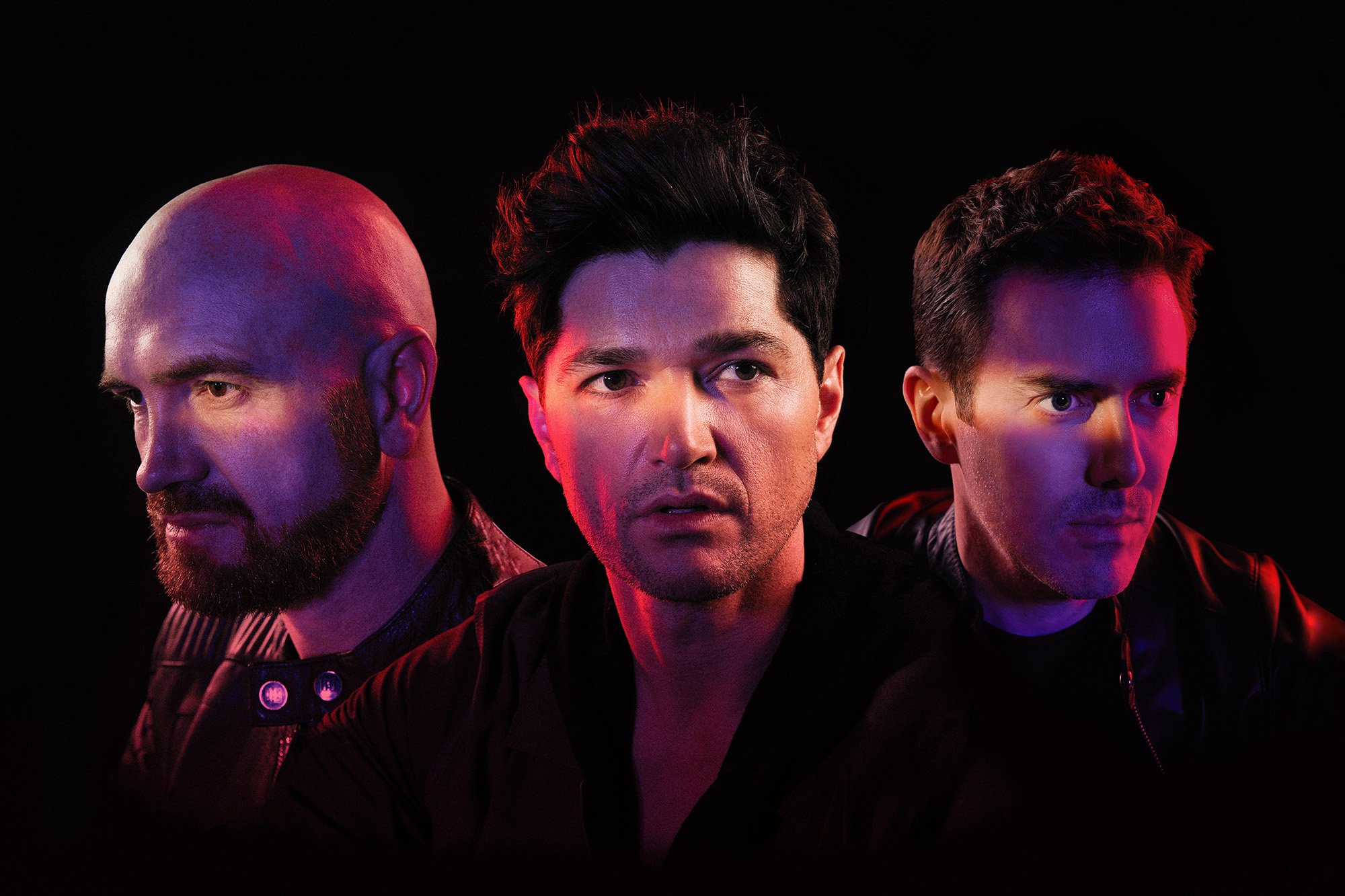 The Script with serious expressions, illuminated by colorful pink and blue lighting, posing against a black background.