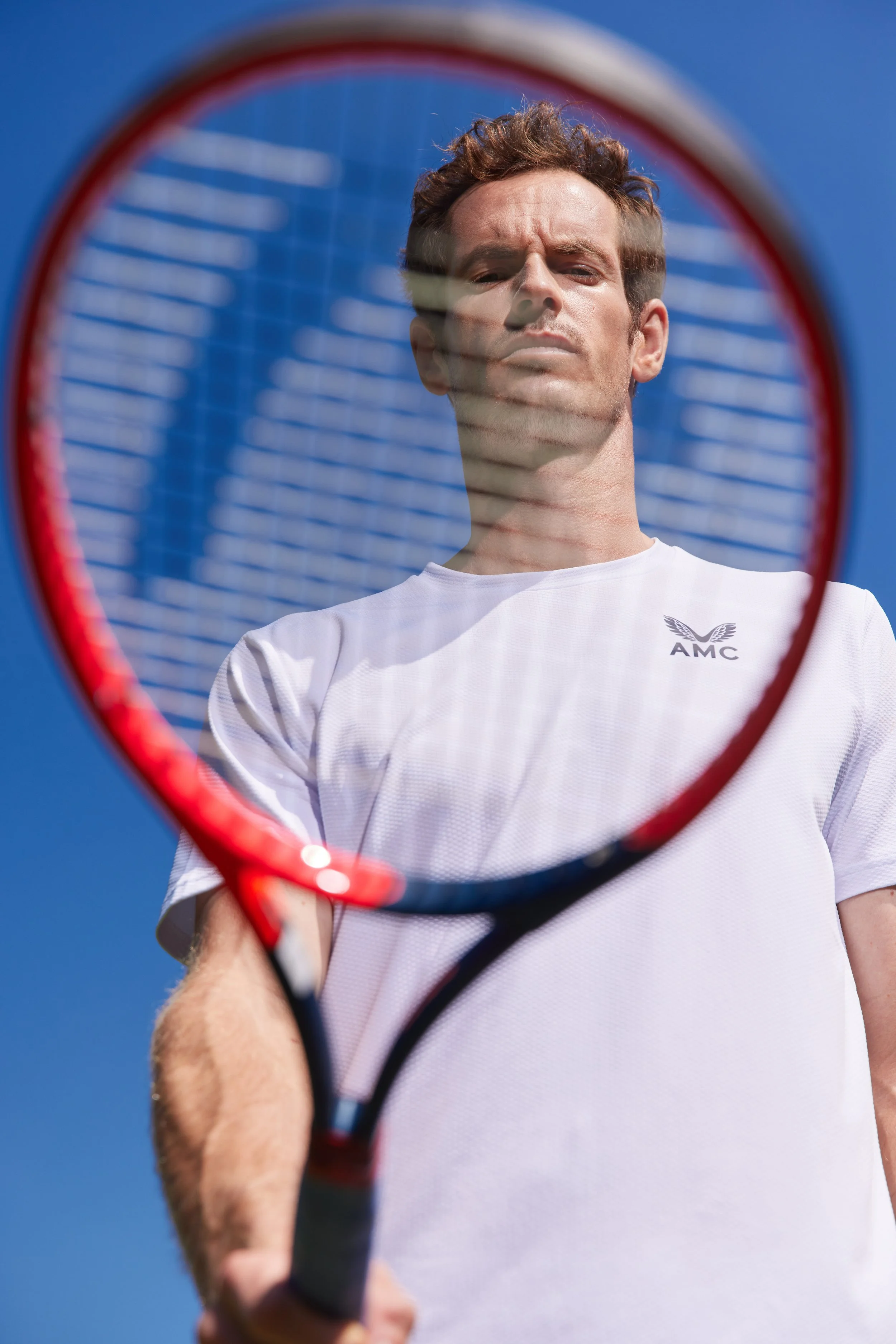 Sir Andy Murray is seen through the strings of a tennis racket, holding the racket towards the camera, with a clear blue sky in the background. He is wearing a white shirt with a logo that says AMC.