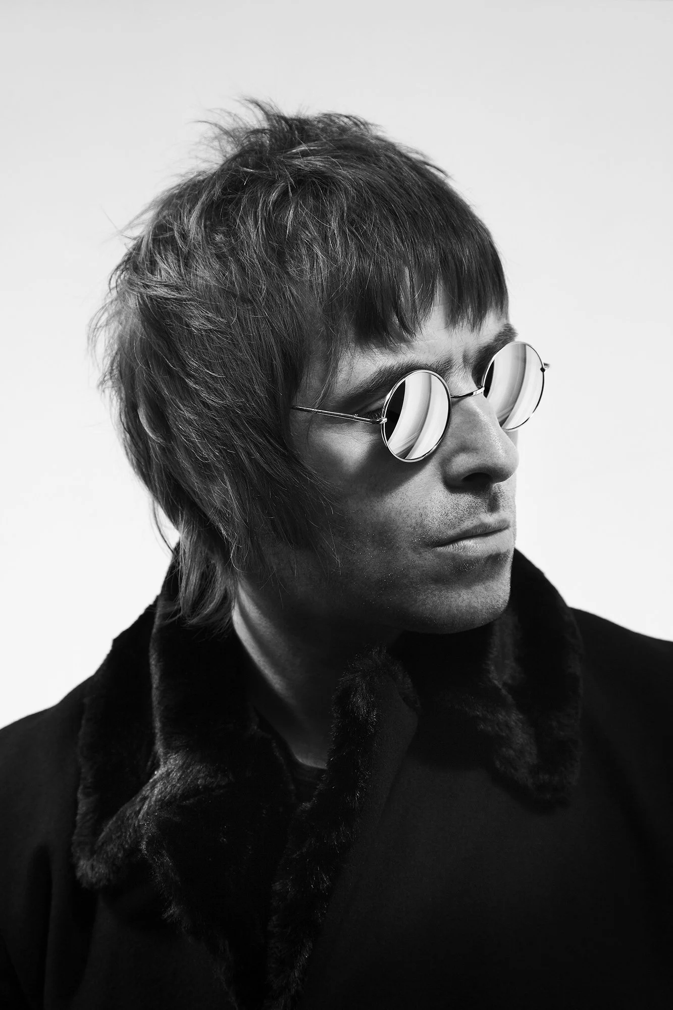 Black and white portrait of Liam Gallagher wearing round sunglasses with striped reflections, medium-length tousled hair, and a dark coat with a fur collar, looking to the side.