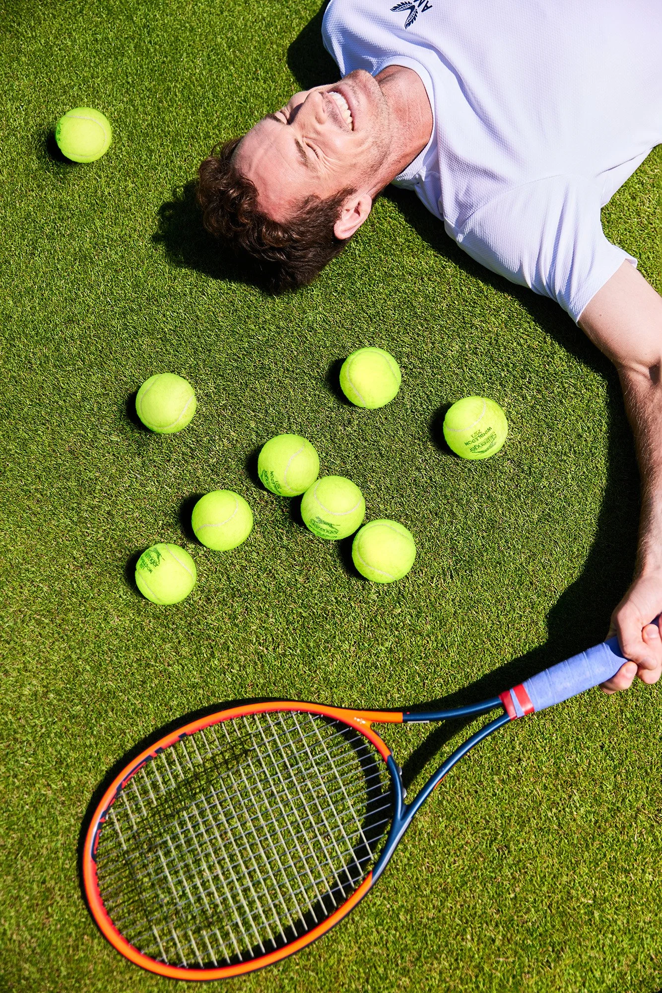 Sir Andy Murray lying on green grass at Wimbledon surrounded by tennis balls and holding a tennis racket, smiling with eyes closed.