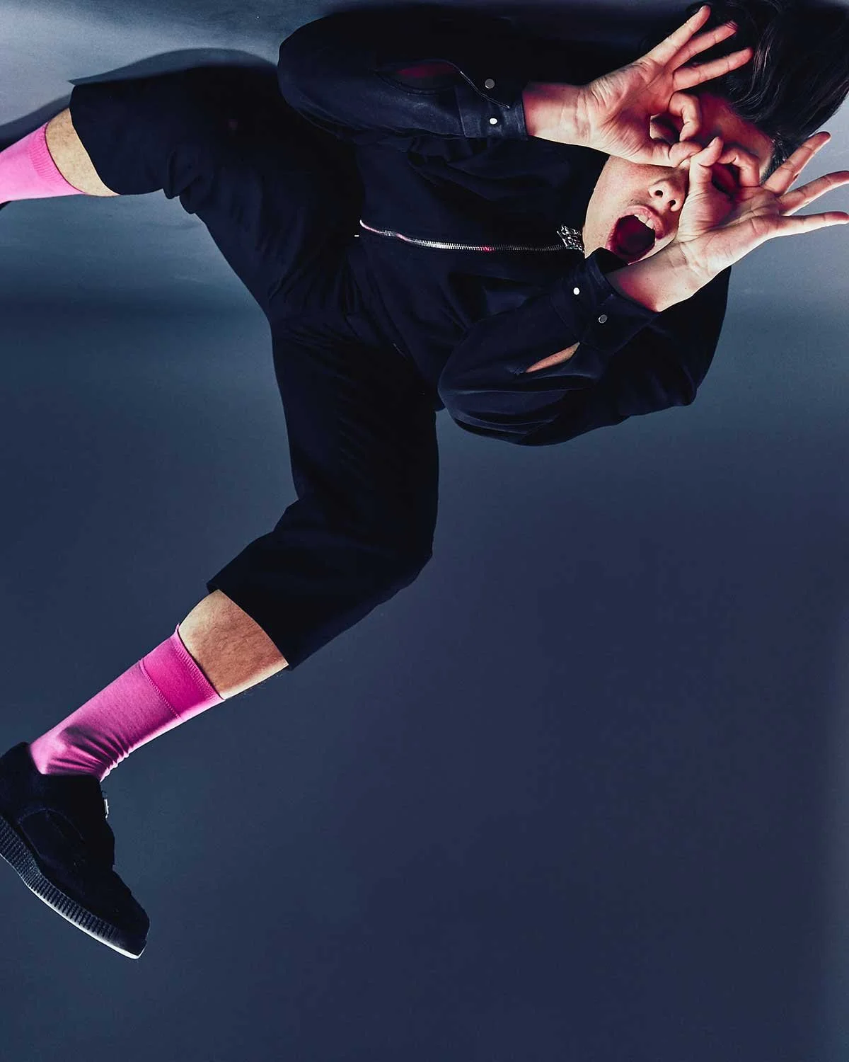Yungblud wearing black clothing with pink socks, making a playful gesture with hands around their eyes, mouth open, against a dark background.