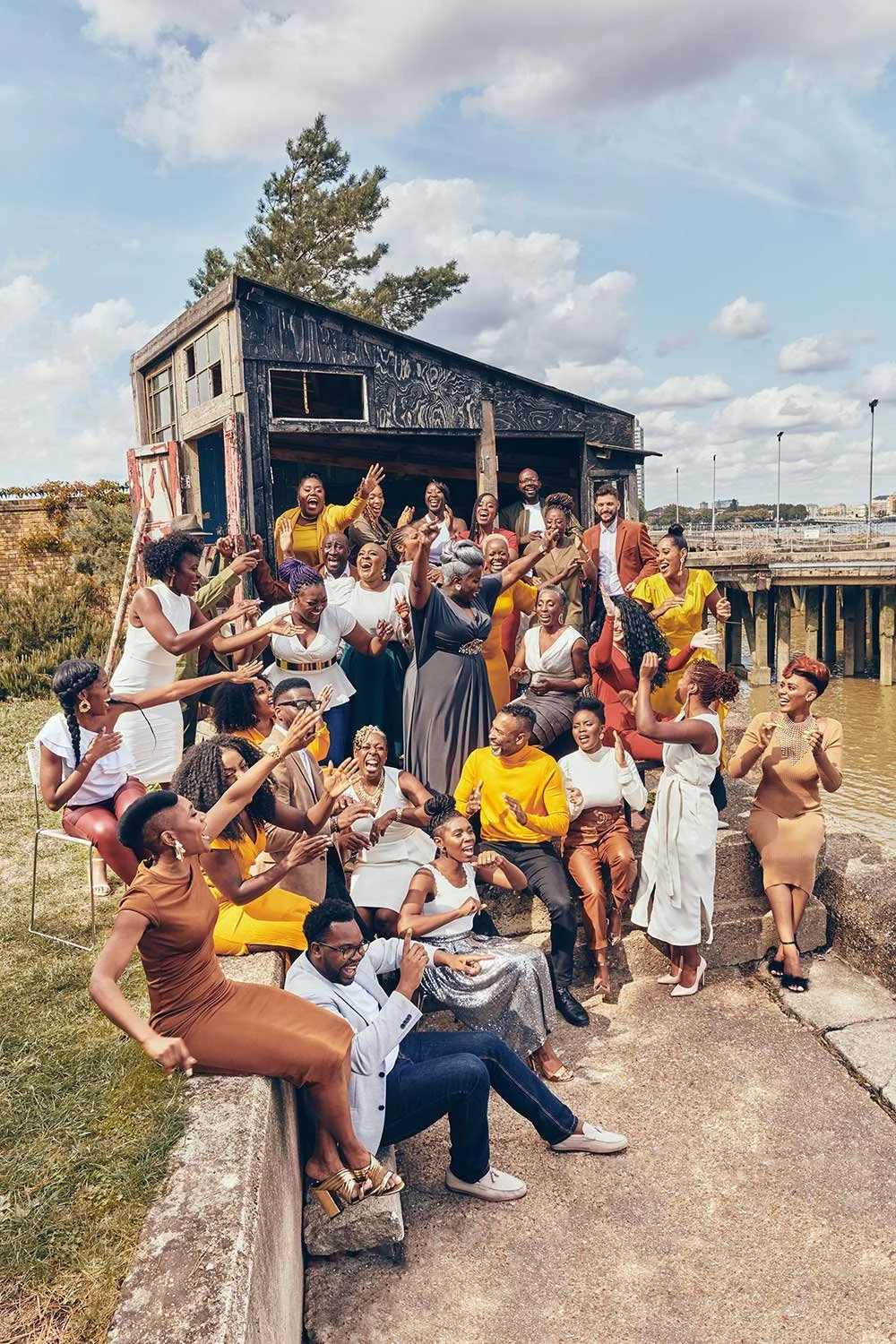 Kingdom Choir celebrating outdoors near a river, with a large, rustic wooden structure in the background. Many are smiling, clapping, and raising their hands, dressed in colorful outfits.