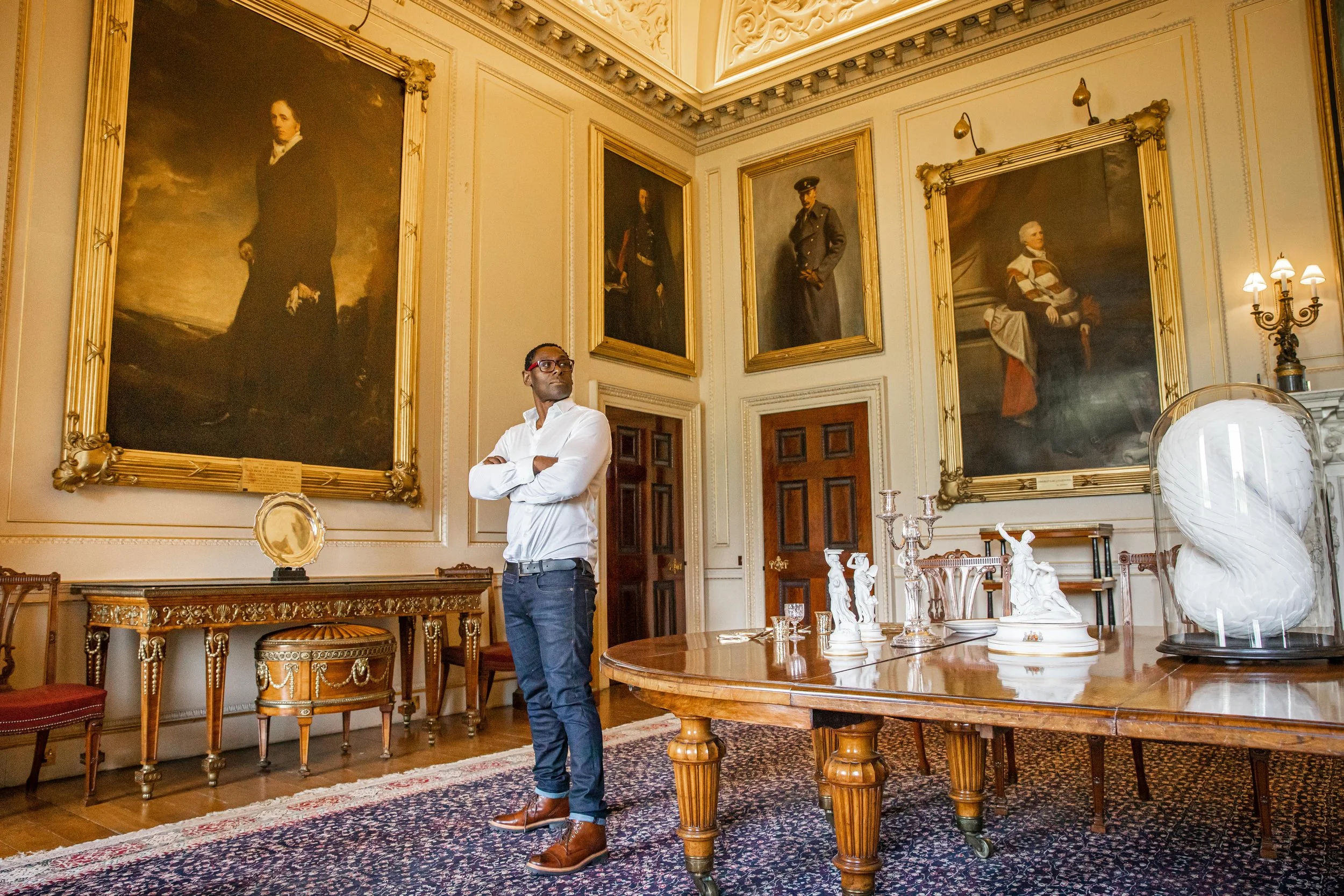 David Harewood in a white shirt and dark pants stands with arms crossed in a grand, ornate room with large, framed classical portraits on the walls featuring his ancestors slave master the Earl of Harewood.
