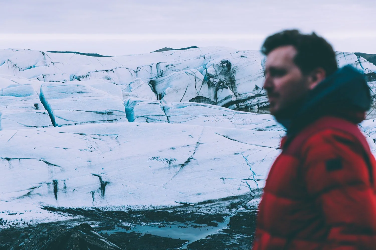 Andrew Whitton wearing a red jacket standing near a glacier with cracked ice and snow-covered terrain in the background in Iceland.