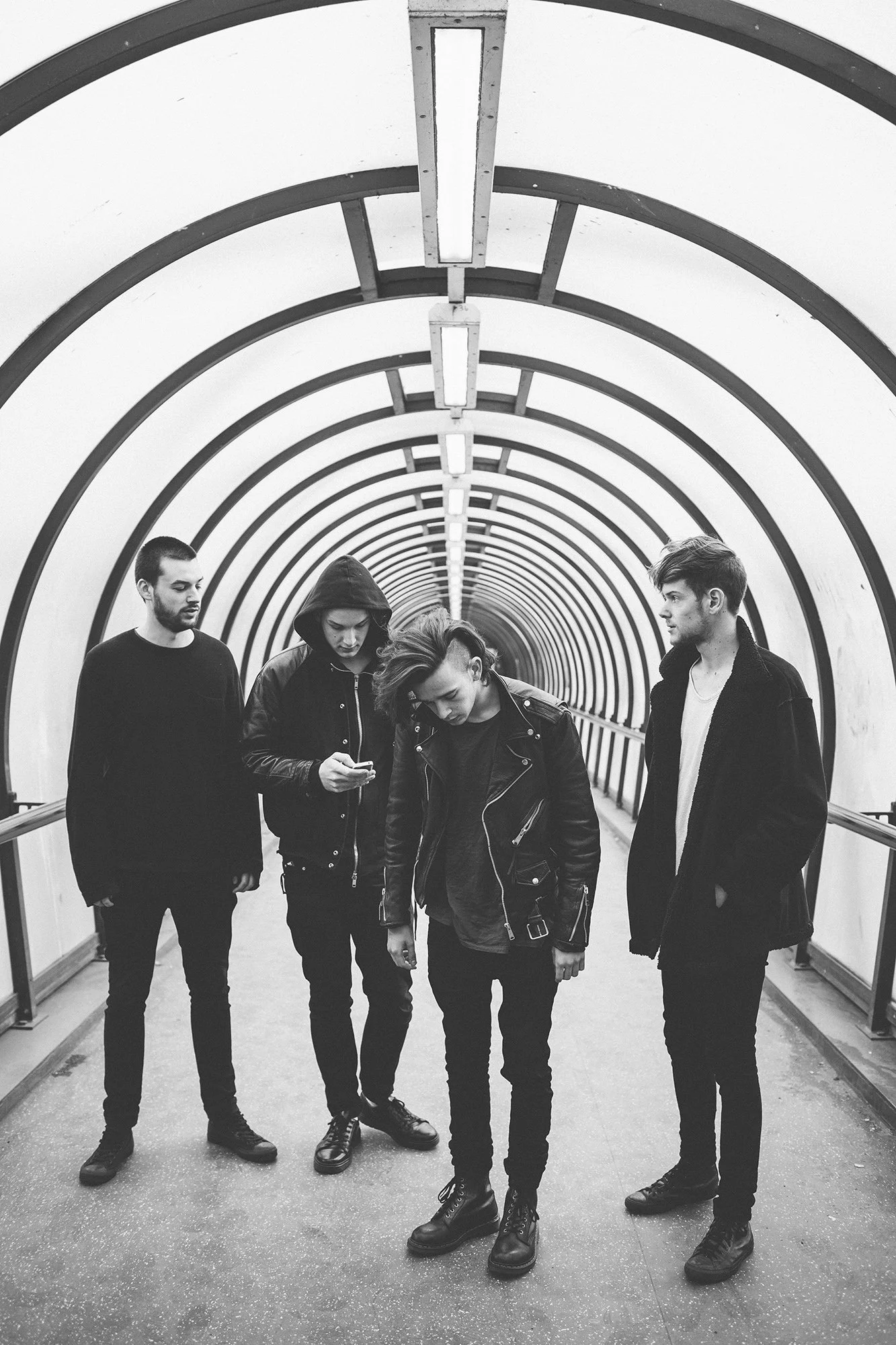 Black-and-white photo of the 1975 standing in an underground pedestrian tunnel with curved arches, some looking down or at their phones, dressed in dark clothing.