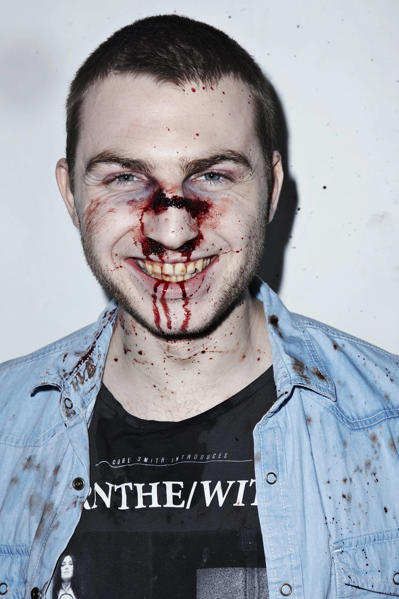 Man with fake blood and makeup simulating a bloody face, smiling at the camera, wearing a black T-shirt and light blue denim shirt.