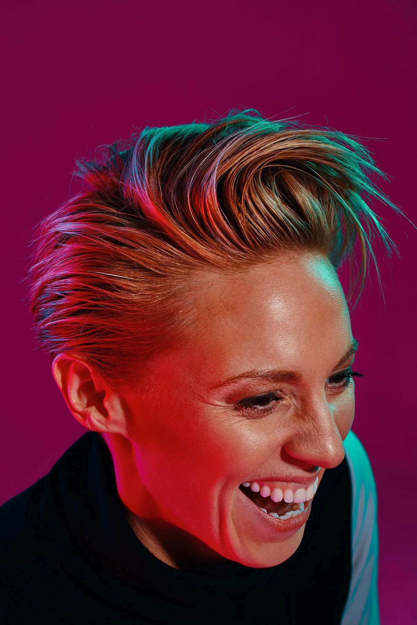 La Roux with a short, platinum blonde hairstyle, laughing and looking to her right, with multicolored lighting highlighting her hair and face.
