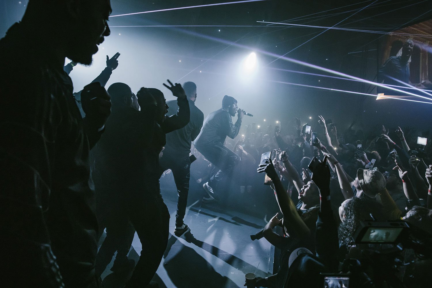 Stormzy crouching and singing into a microphone on stage at a concert, surrounded by enthusiastic audience members reaching out and taking photos, with bright stage lights and laser beams illuminating the scene. 