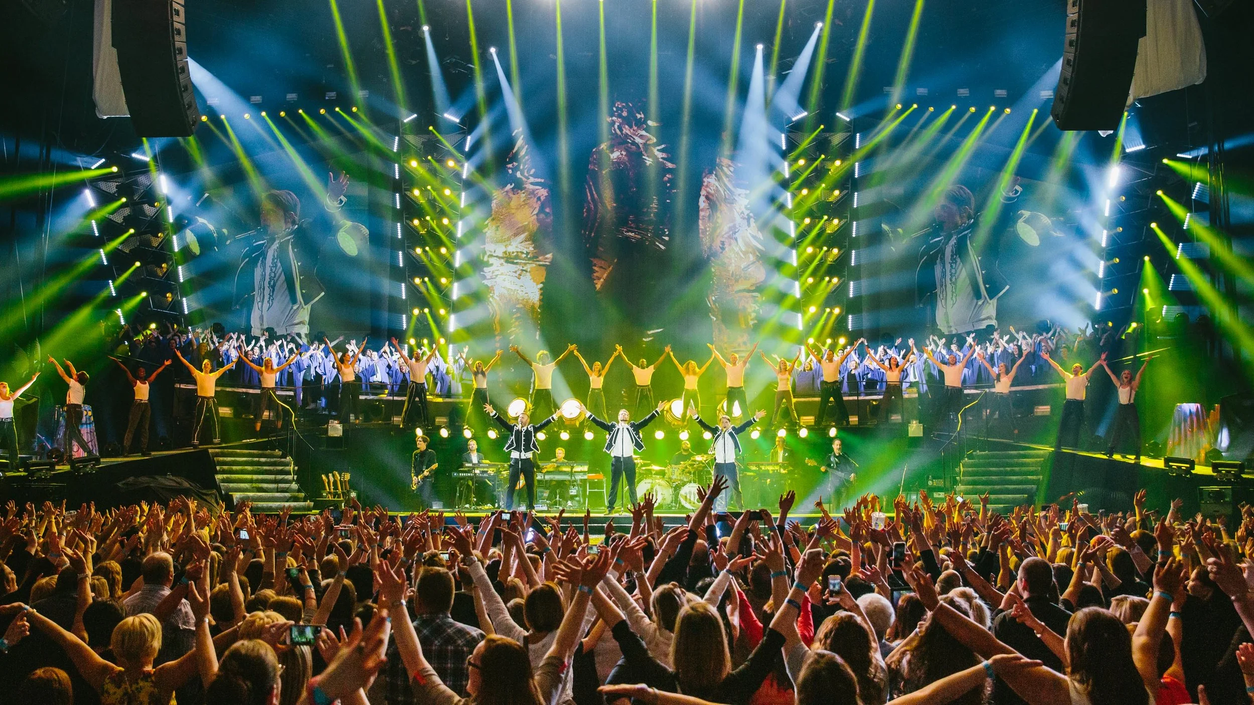 A lively Take That concert scene with a stage full of performers, dancers, and musicians surrounded by colorful lights, with an enthusiastic audience cheering and raising their hands.