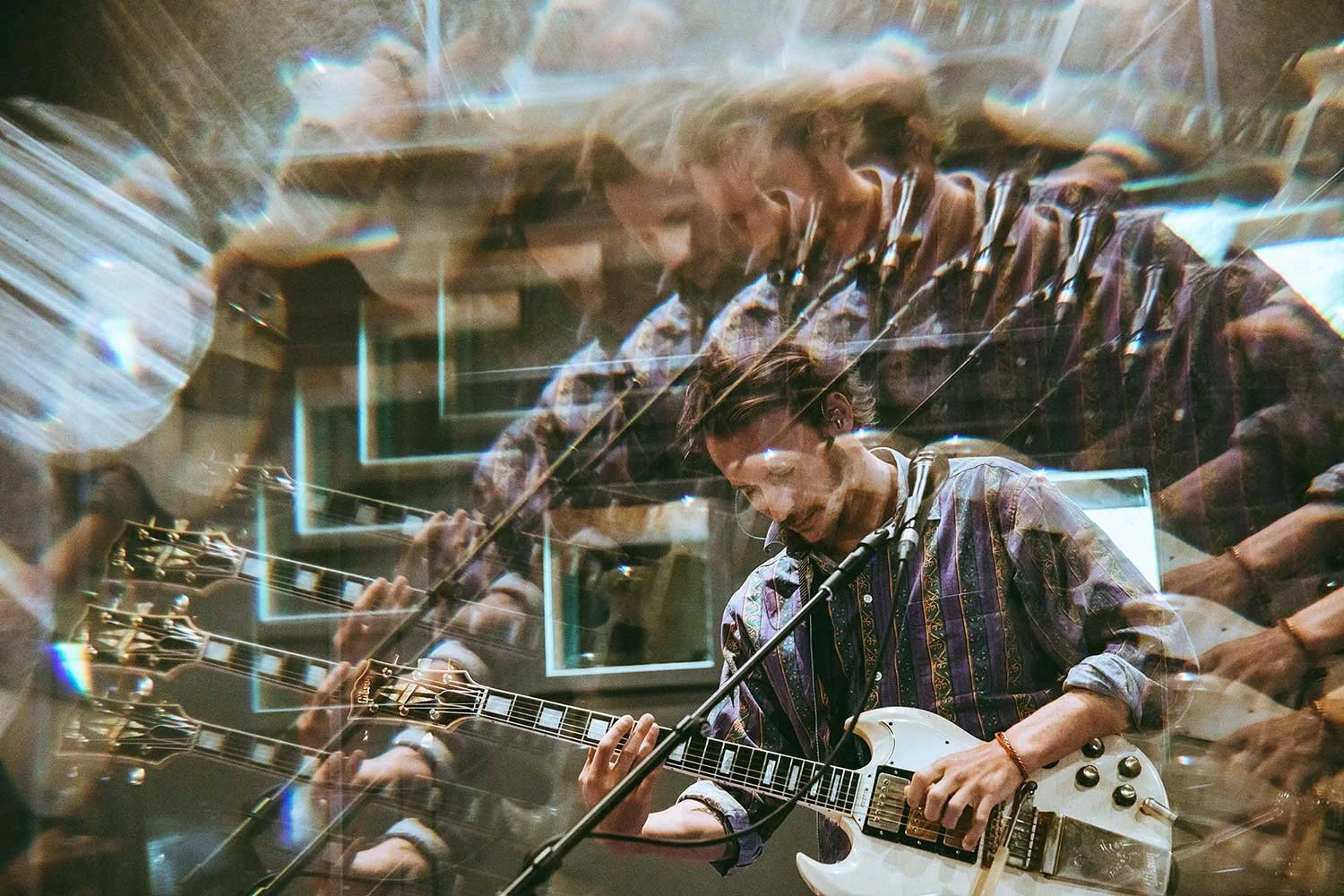 A woman playing an electric guitar, seen through a reflective and layered glass surface, creating multiple overlapping reflections of her and the guitar.