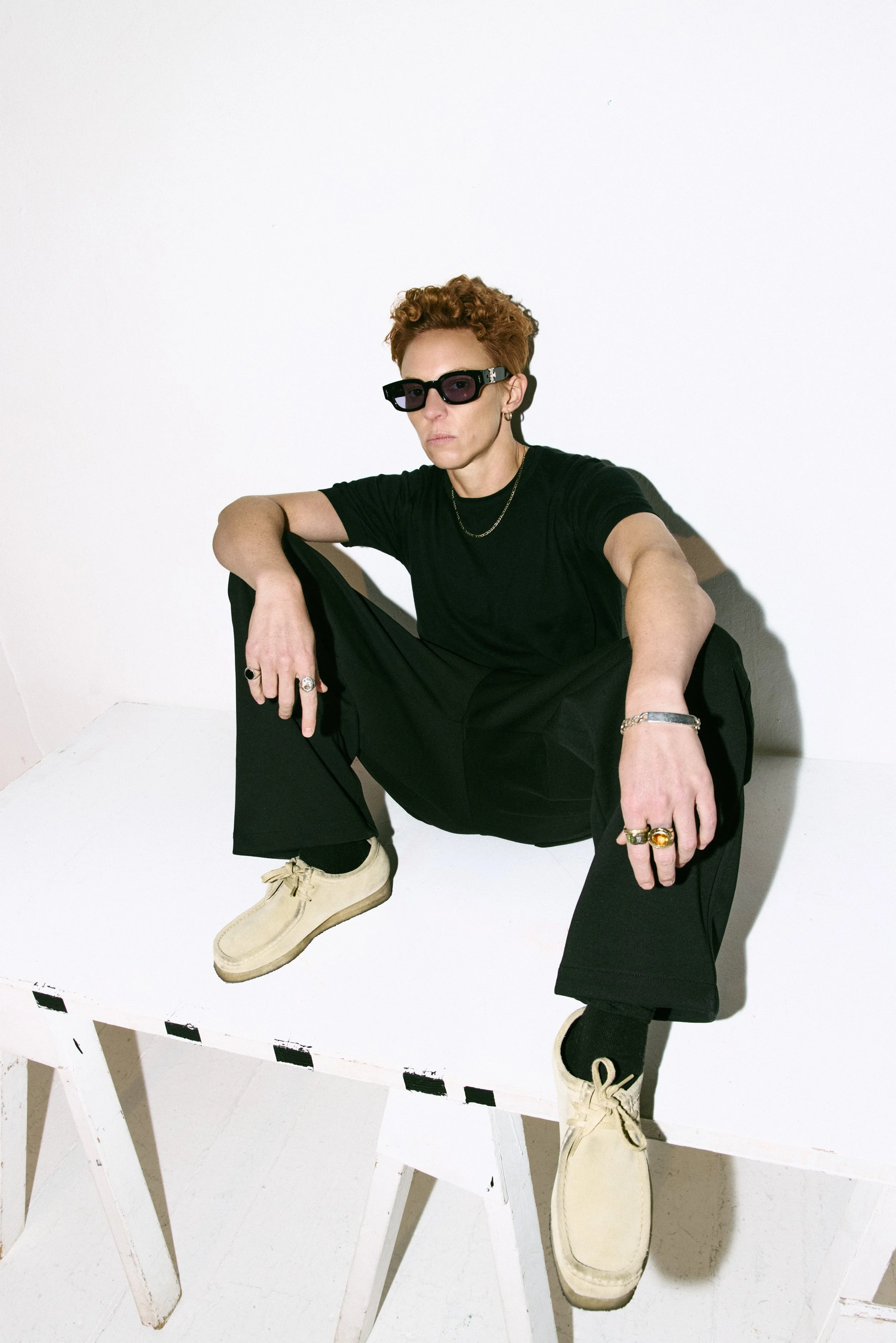 La Roux with short curly red hair wearing sunglasses, black shirt, black pants, and cream-colored shoes, sitting on a white wooden bench against a white wall.