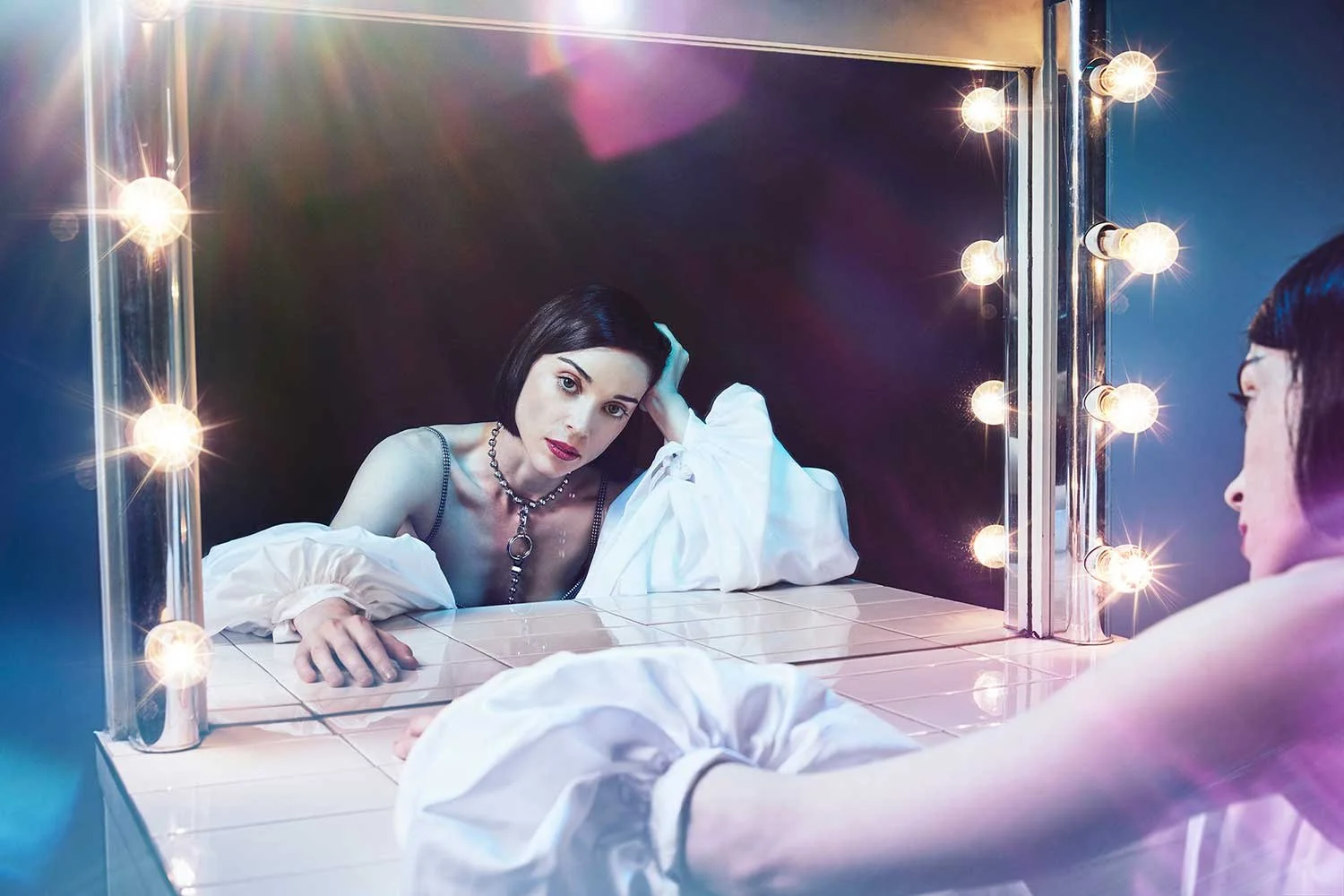 St Vincent with short black hair and dark lipstick looking into a mirror with glowing light bulbs, resting her head on her hand, in a dressing room