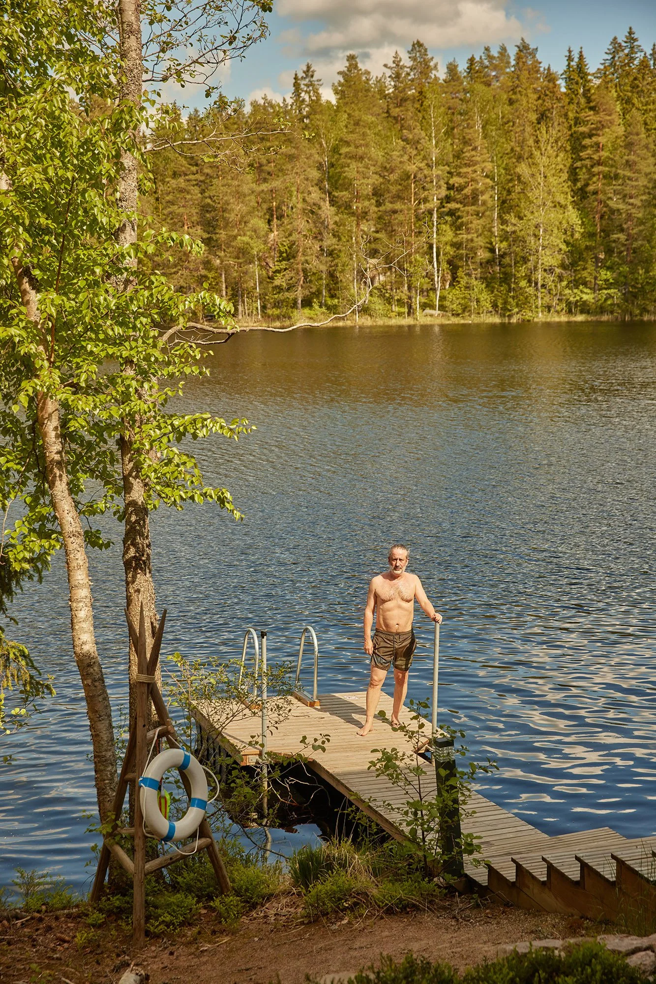 David Baddiel on a wooden dock by a lake, surrounded by trees, holding a pole, with a life preserver hanging on a nearby structure in Finland.