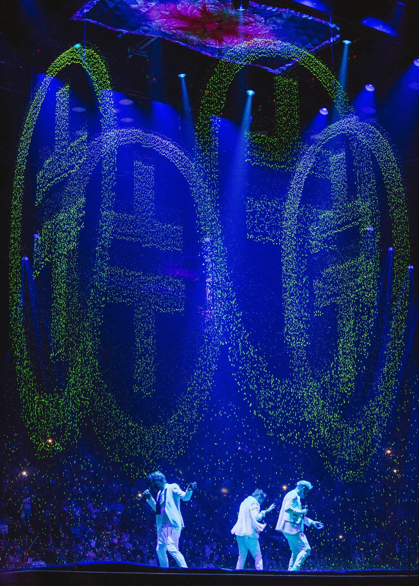 Take That large illuminated display with the words 'LOVE' and 'PEACE' written in bright, colorful lights, with three performers in white suits dancing on stage beneath the display.