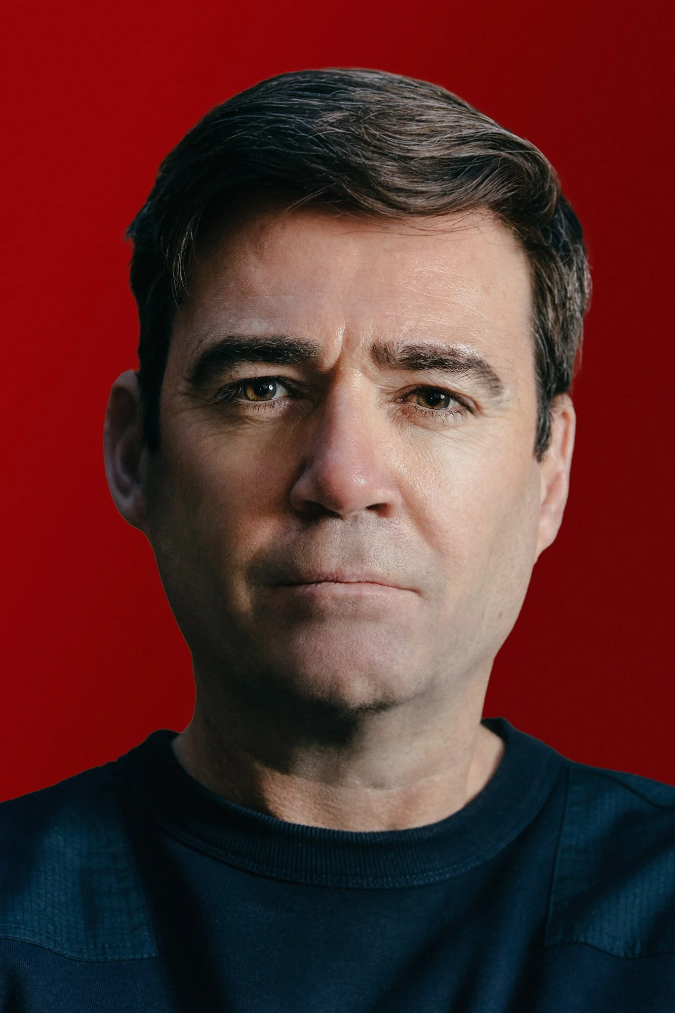 A close-up portrait of Andy Burnham with dark hair, blue eyes, and wearing a navy shirt, against a red background.