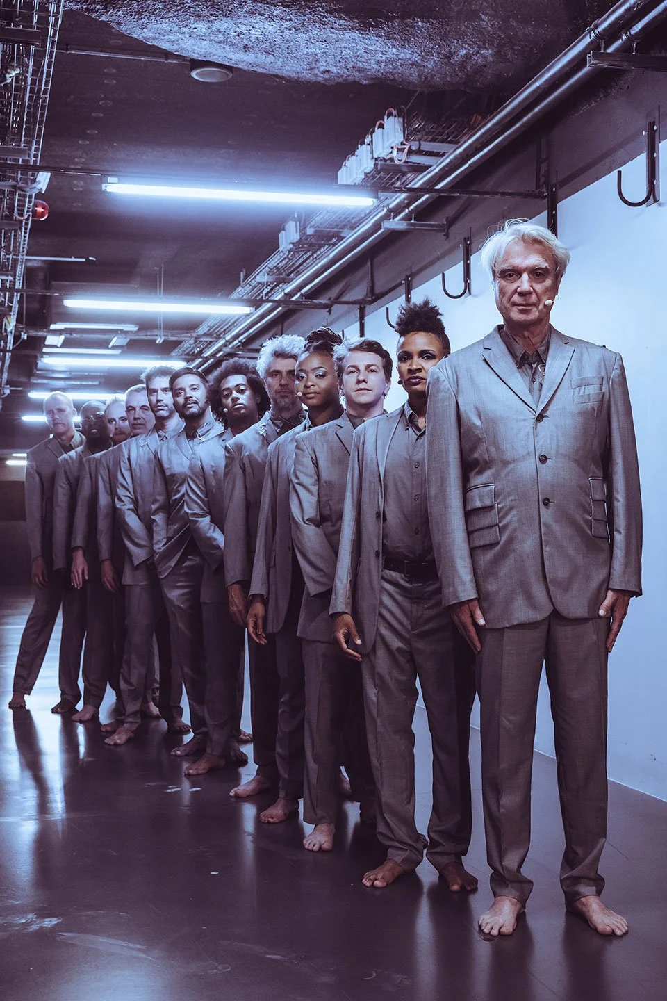 David Byrne and his band in grey suits standing in a line indoors under fluorescent lights, with some barefoot, facing forward with serious expressions. Pre performance