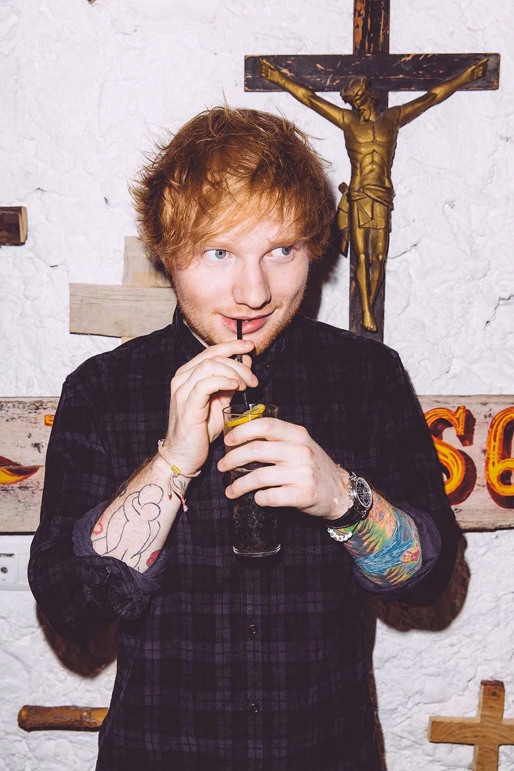 Ed Sheeran with red hair and tattoos sipping a drink through a straw, standing in front of a crucifix on a white textured wall.