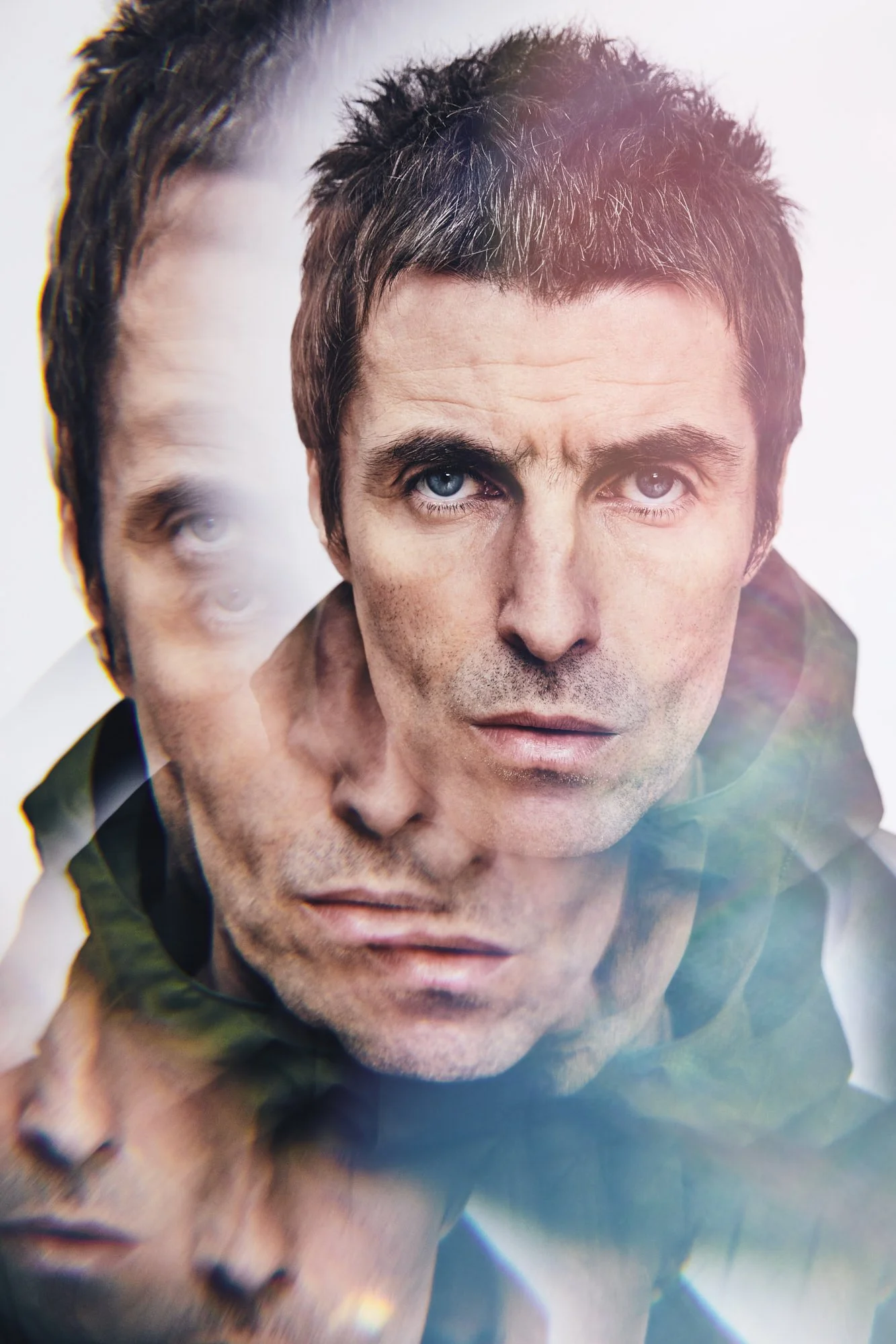 Close-up of Liam Gallagher with dark hair, blue eyes, and stubble, looking intently at the camera. There is a superimposed, semi-transparent image of the same man in the background.