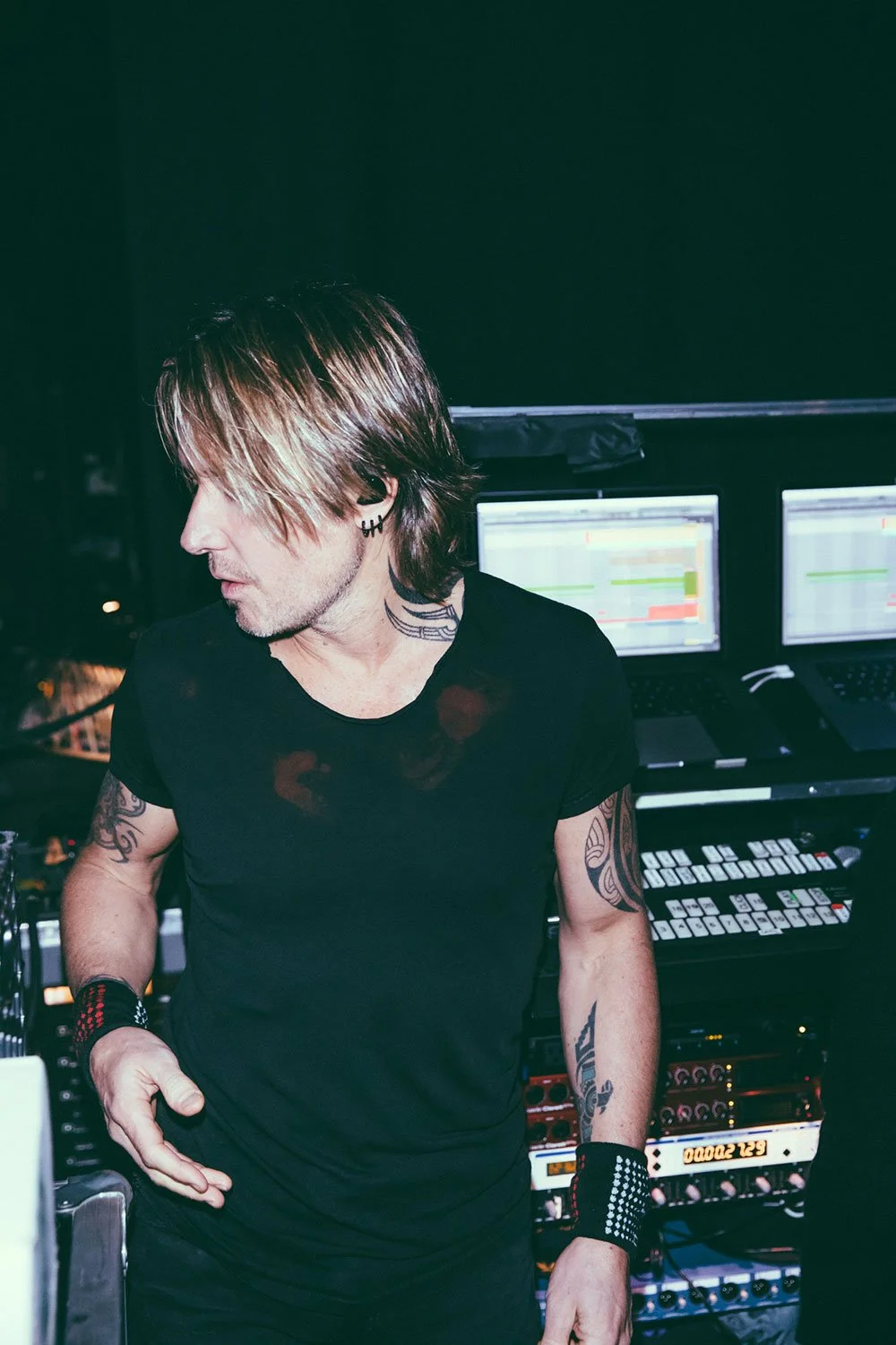Music artist, Keith Urban with tattoos, wearing black shirt and accessories, working with music equipment and two monitors in a dimly lit setting.