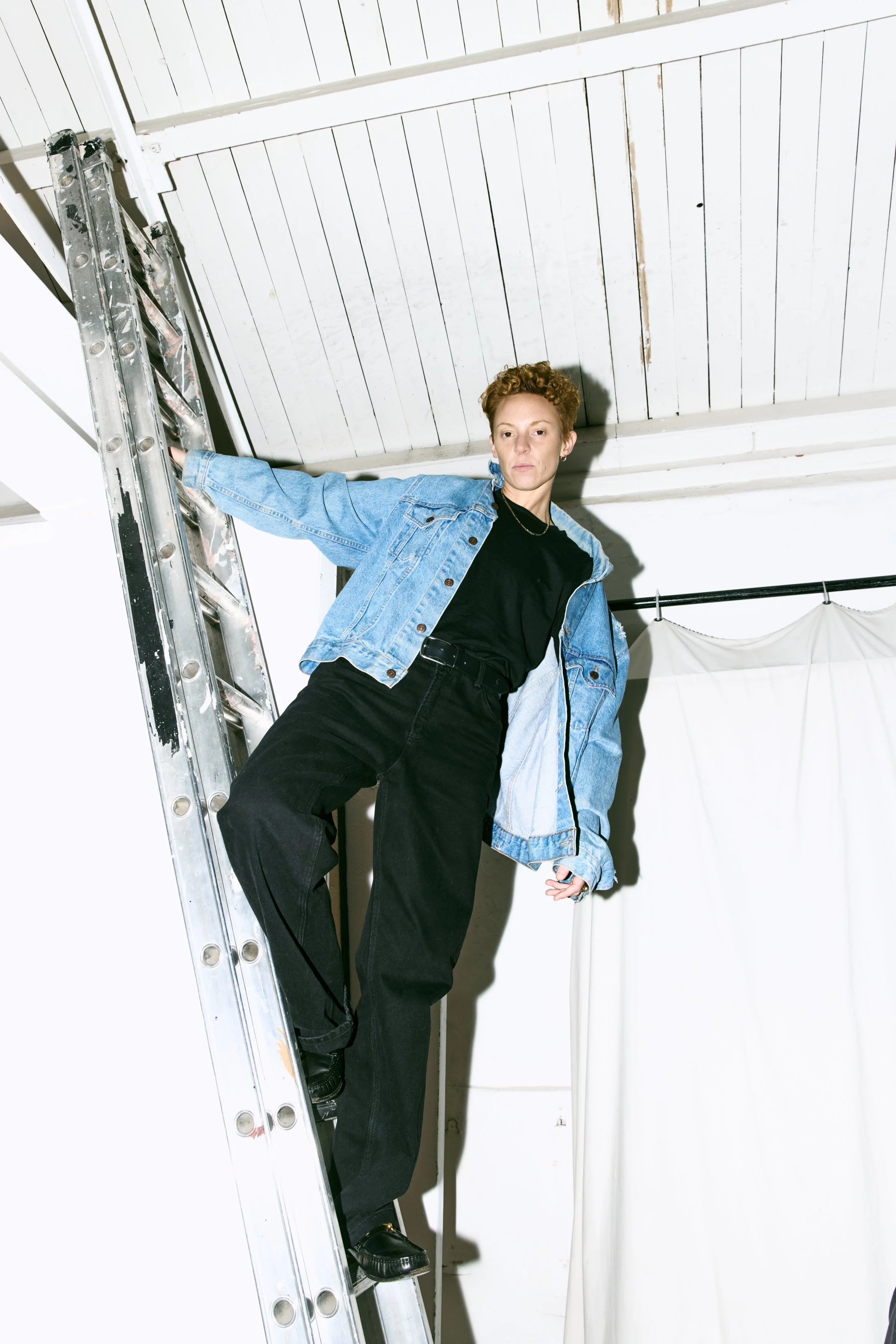La Roux with short curly hair wearing a black shirt and black pants, with a blue denim jacket, standing on a ladder in a white room, with a curtain hanging in the background.
