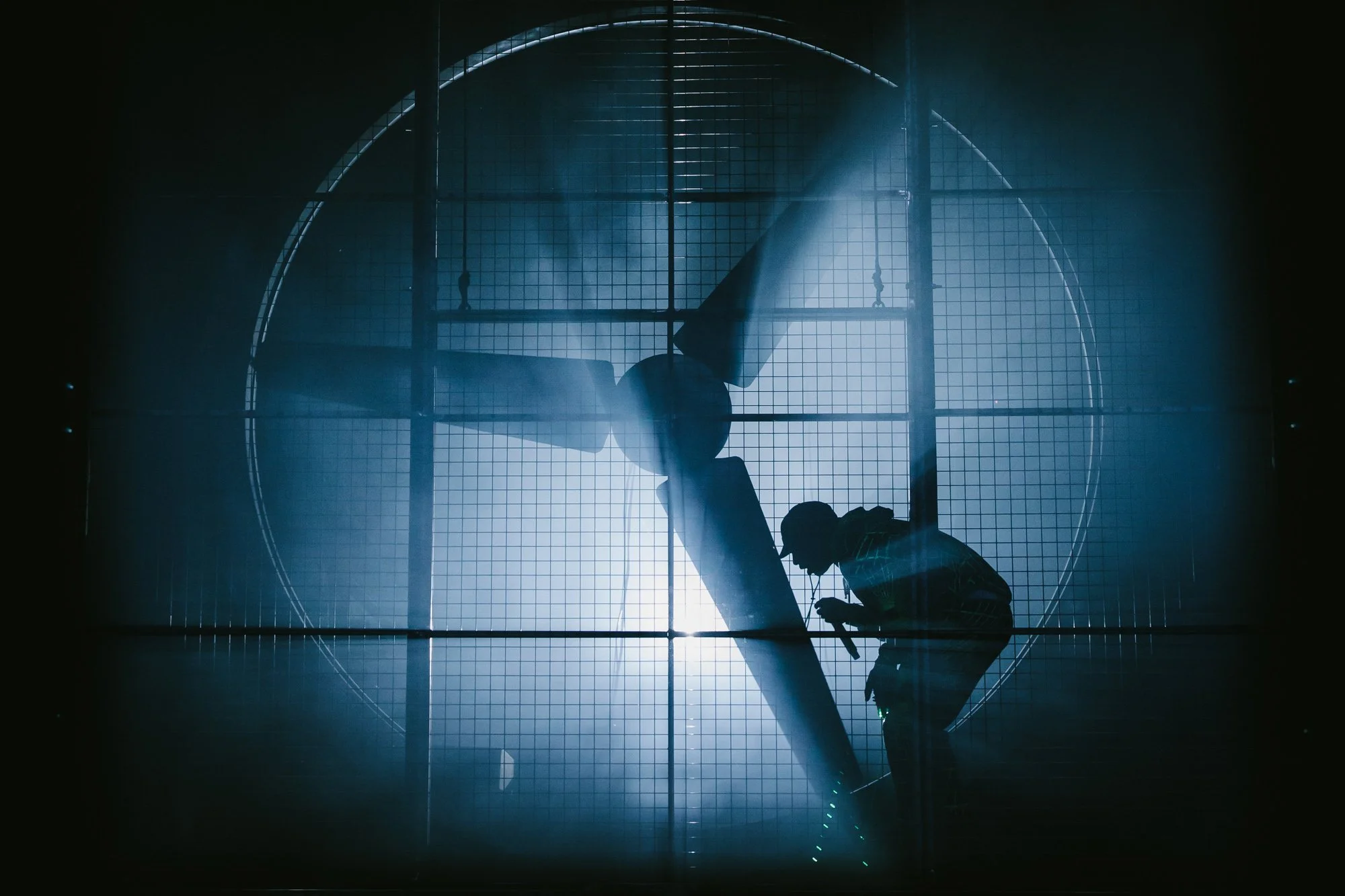 Silhouette of Skepta performing maintenance or inspection on a large industrial fan or turbine behind a wire mesh or grill, with bright light creating a dramatic effect.