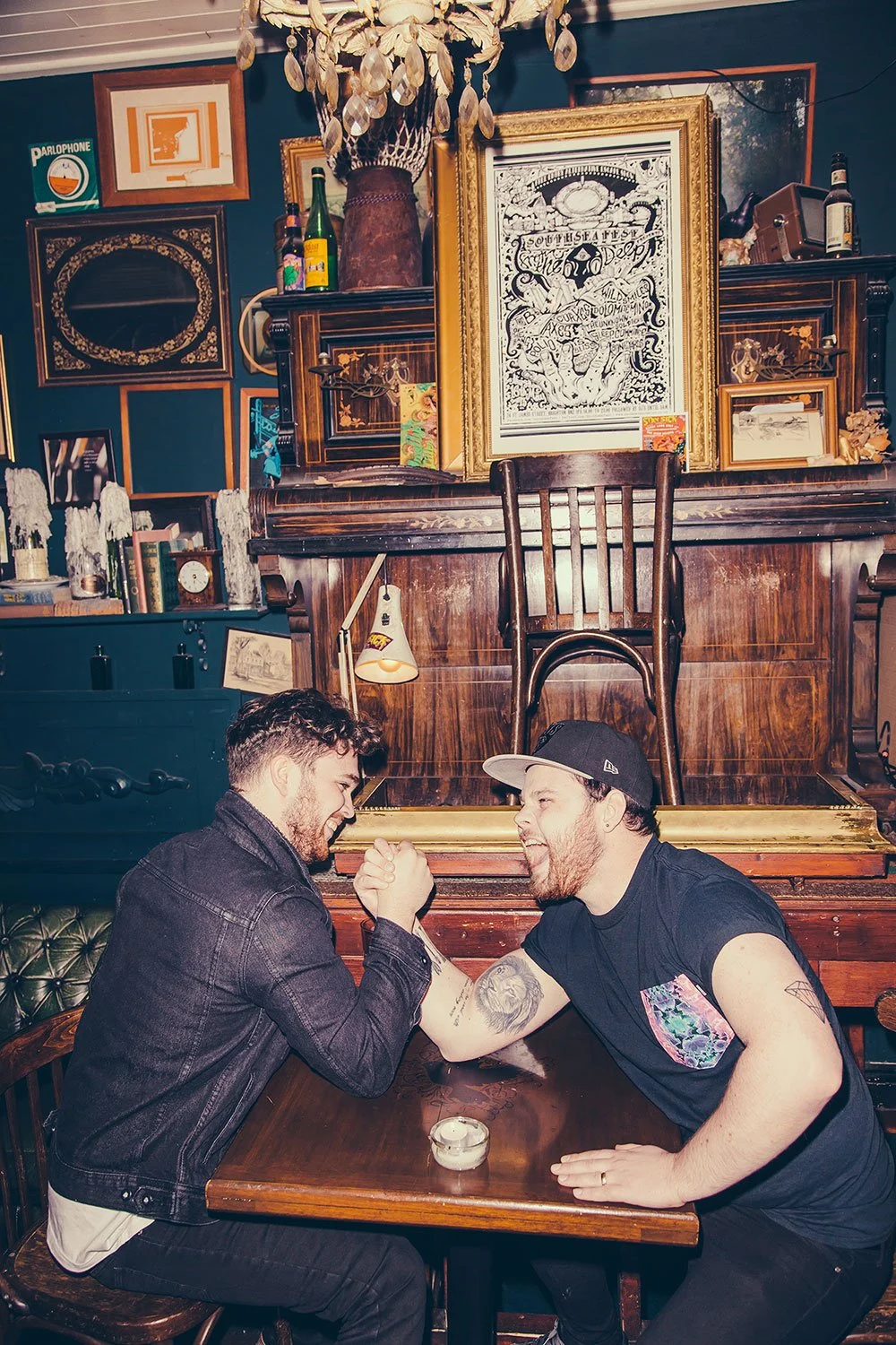 Royal Blood arm wrestling at a wooden table in a cozy, vintage-style room with artwork and bottles on the shelf behind them.