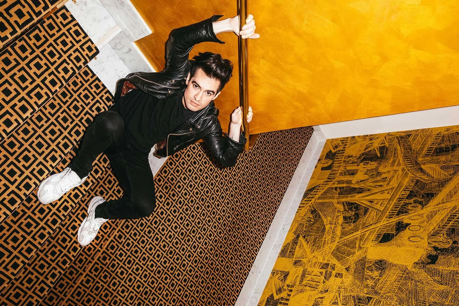 Brendon Irie of Panic at the disco, dressed in a black leather jacket and black pants, is lying on a patterned carpeted floor, holding onto a wooden railing and looking at the camera. 