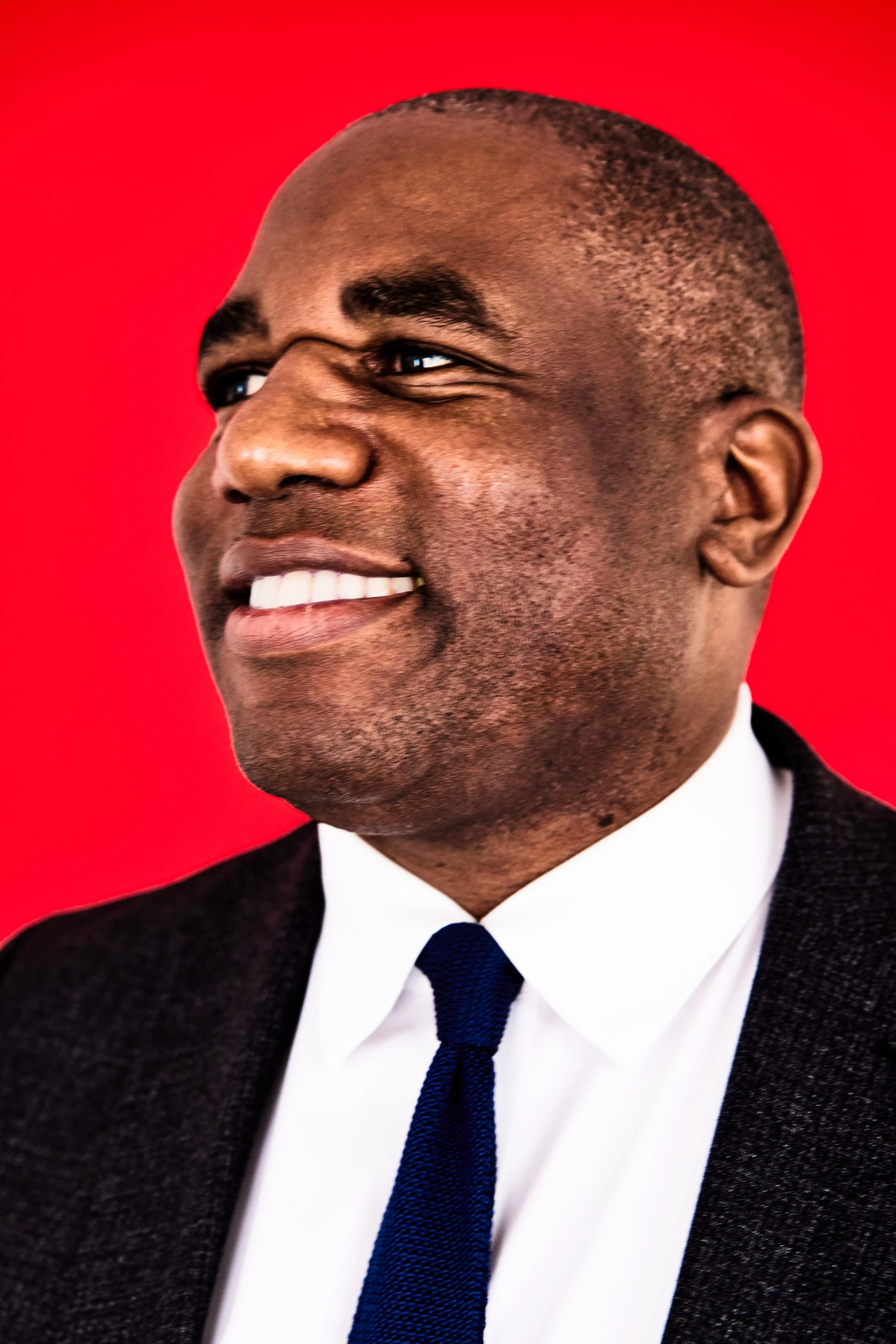 Portrait of a smiling David Lammy MP wearing a dark suit, white shirt, and blue tie, against a red background.